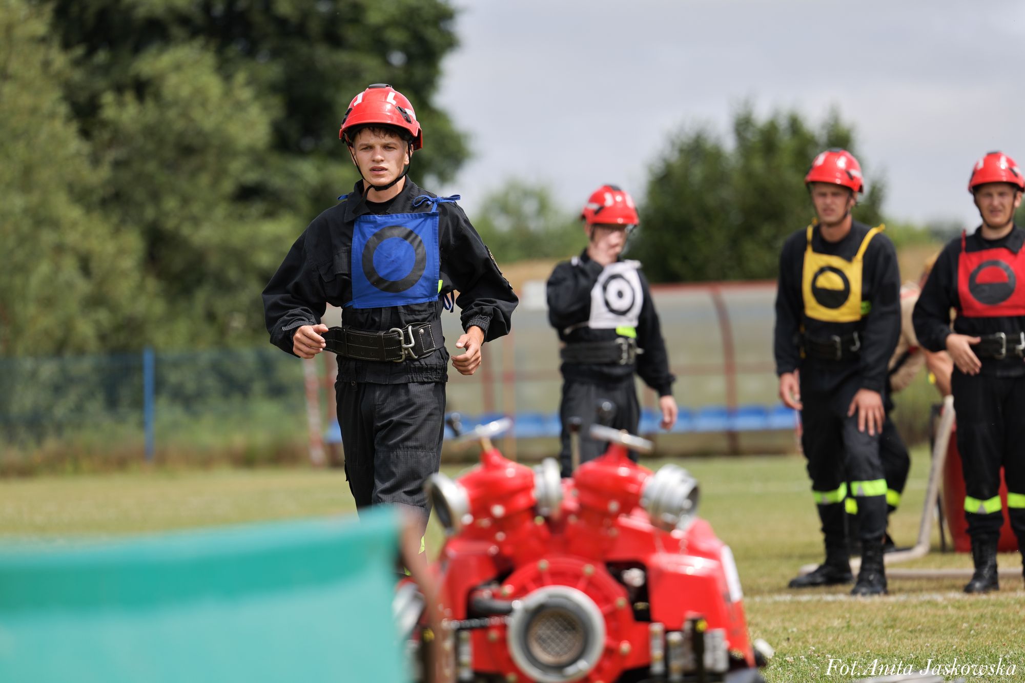 Czterech mężczyzn w czarnych mundurach i czerwonych hełmach, każdy z kolorową kamizelką z symbolem, stoją na trawie, na pierwszym planie czerwony sprzęt strażacki.