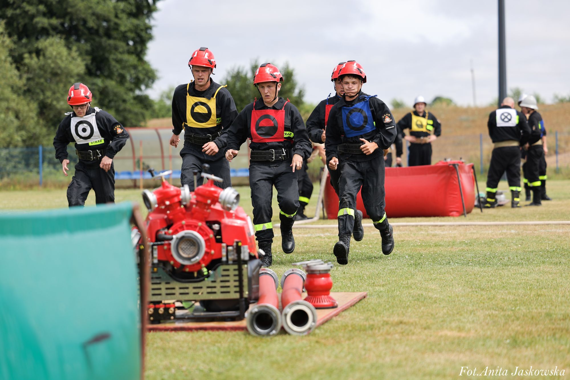 Grupa strażaków w kaskach i kolorowych kamizelkach biegnie po trawniku obok sprzętu pożarniczego i węży.