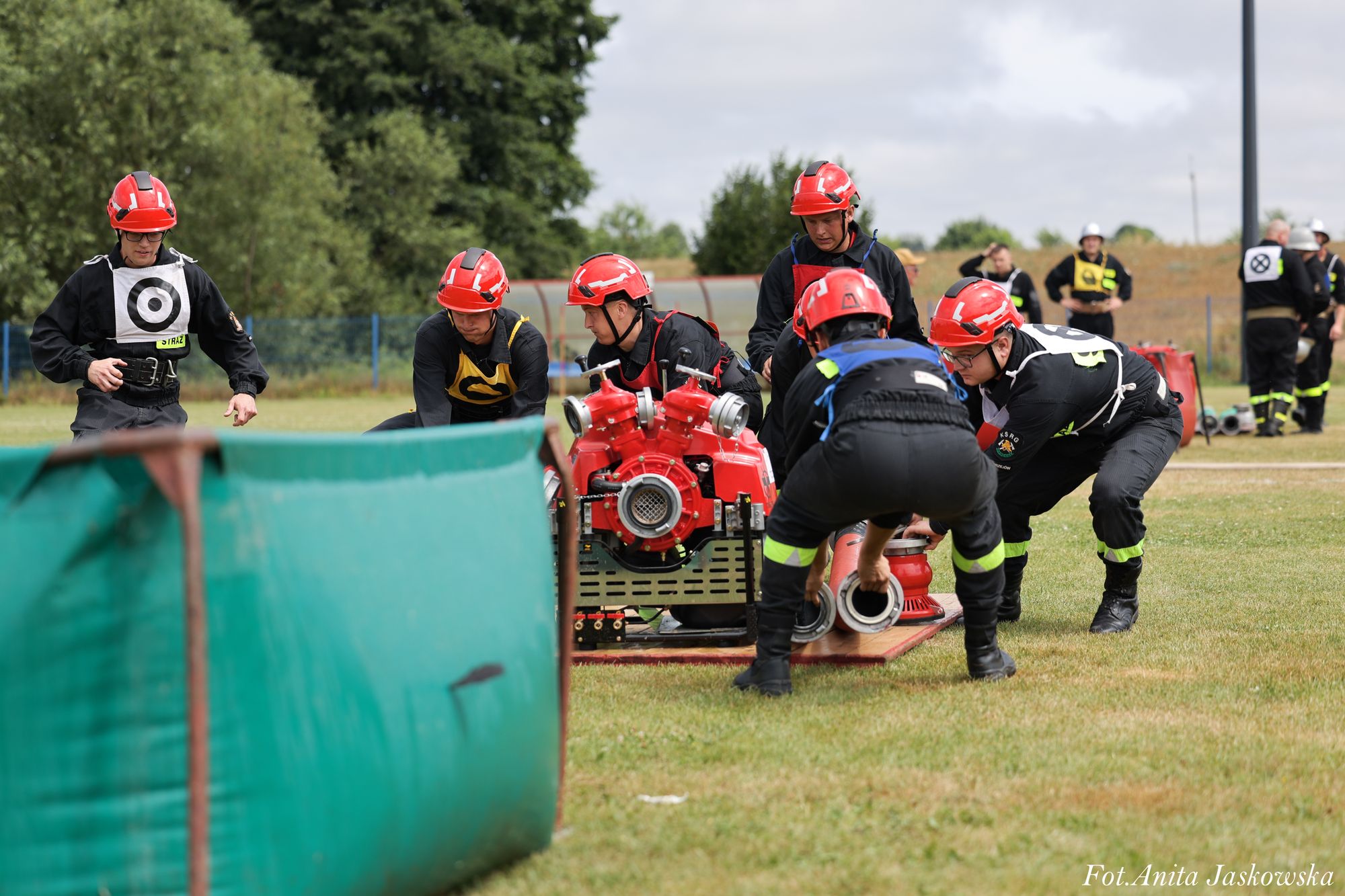 Grupa strażaków w czarnych mundurach i czerwonych hełmach obsługuje czerwoną pompę położoną na trawie, w tle zieleń i inni strażacy.