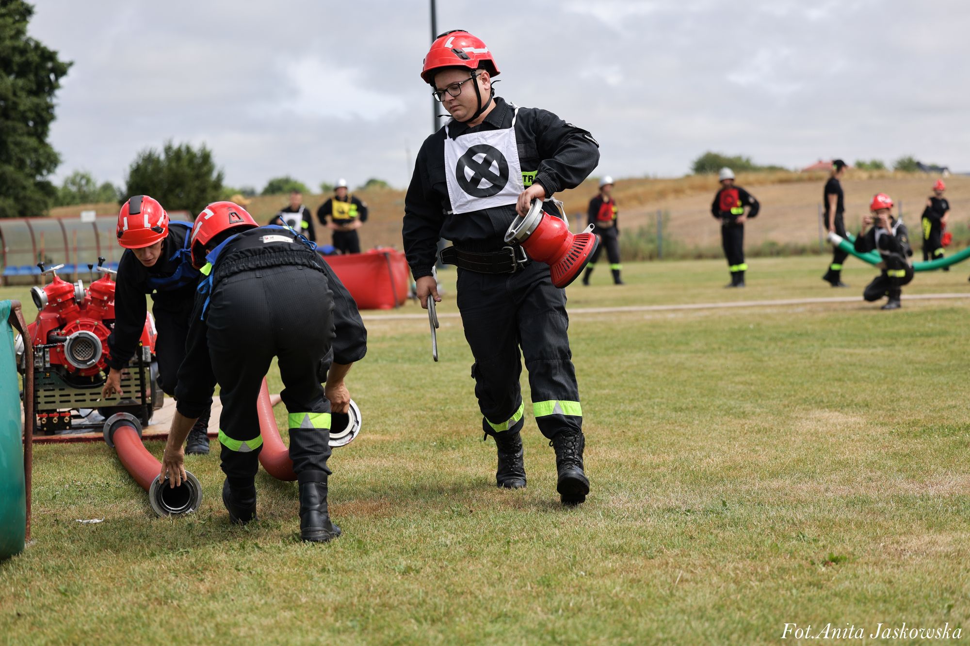Strażacy w czarnych mundurach z odblaskowymi paskami i czerwonych kaskach rozkładają czerwone węże strażackie na zielonym polu trawiastym.