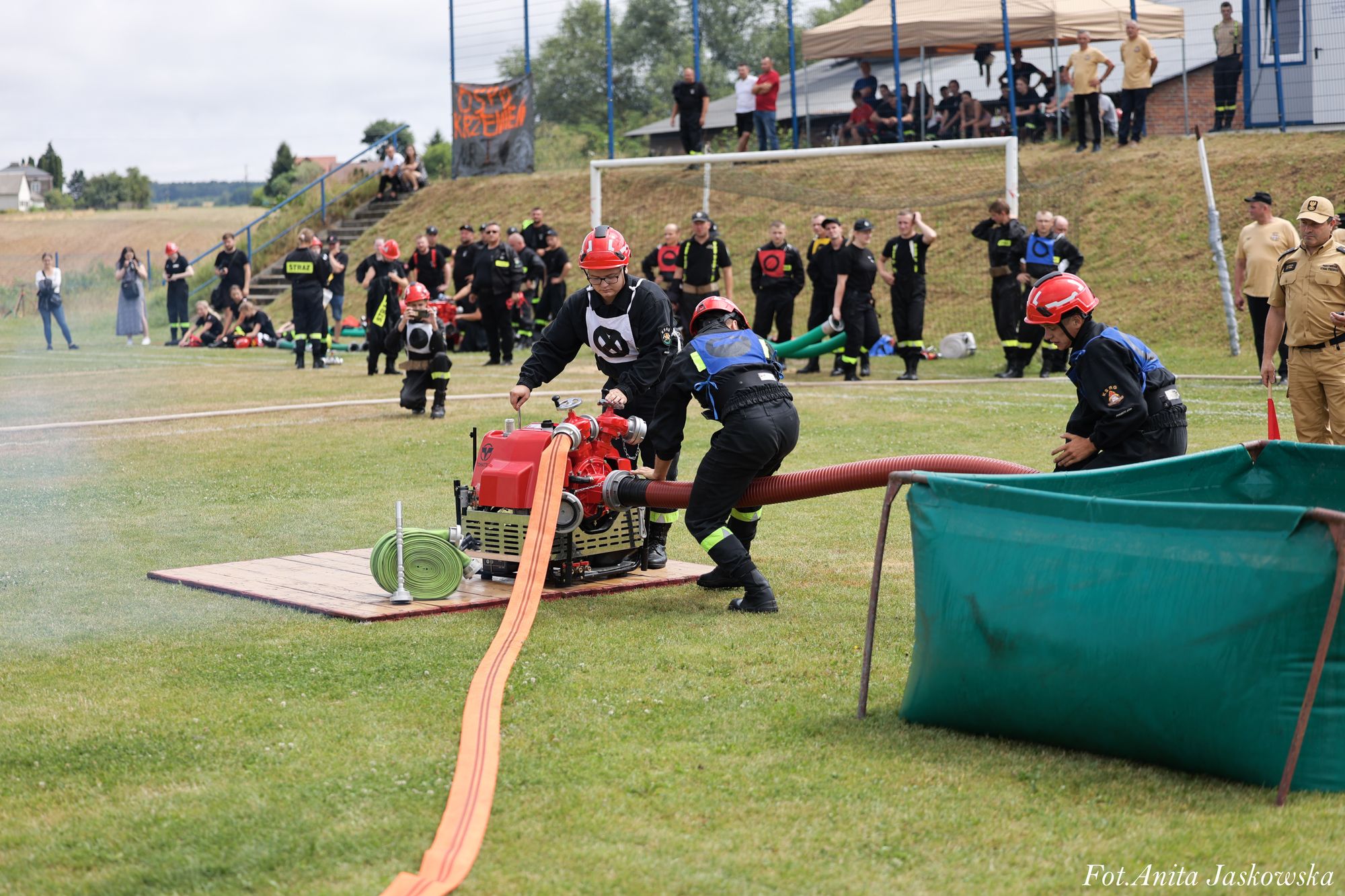 Trzech strażaków w czarnych mundurach i czerwonych kaskach obsługuje czerwony sprzęt połączony z zielonym zbiornikiem i pomarańczowym wężem na zielonej trawie, w tle grupa osób i bramka.