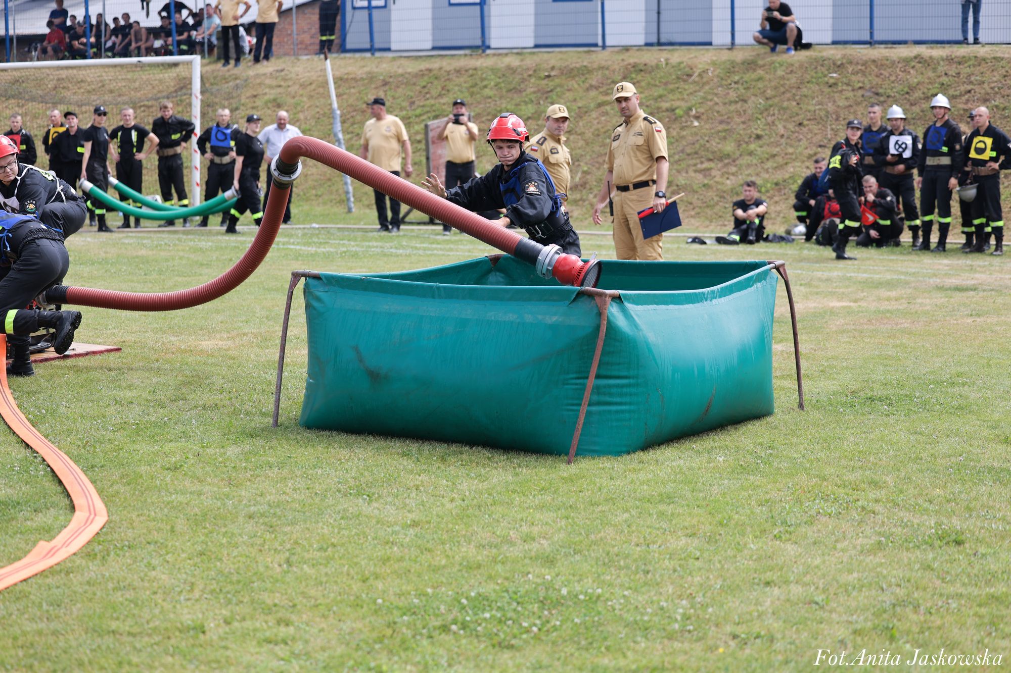 Strażacy w czarnych ubraniach i czerwonych hełmach przy zielonym zbiorniku na wodę na trawniku, w tle osoby w beżowych mundurach i widzowie na skarpie.