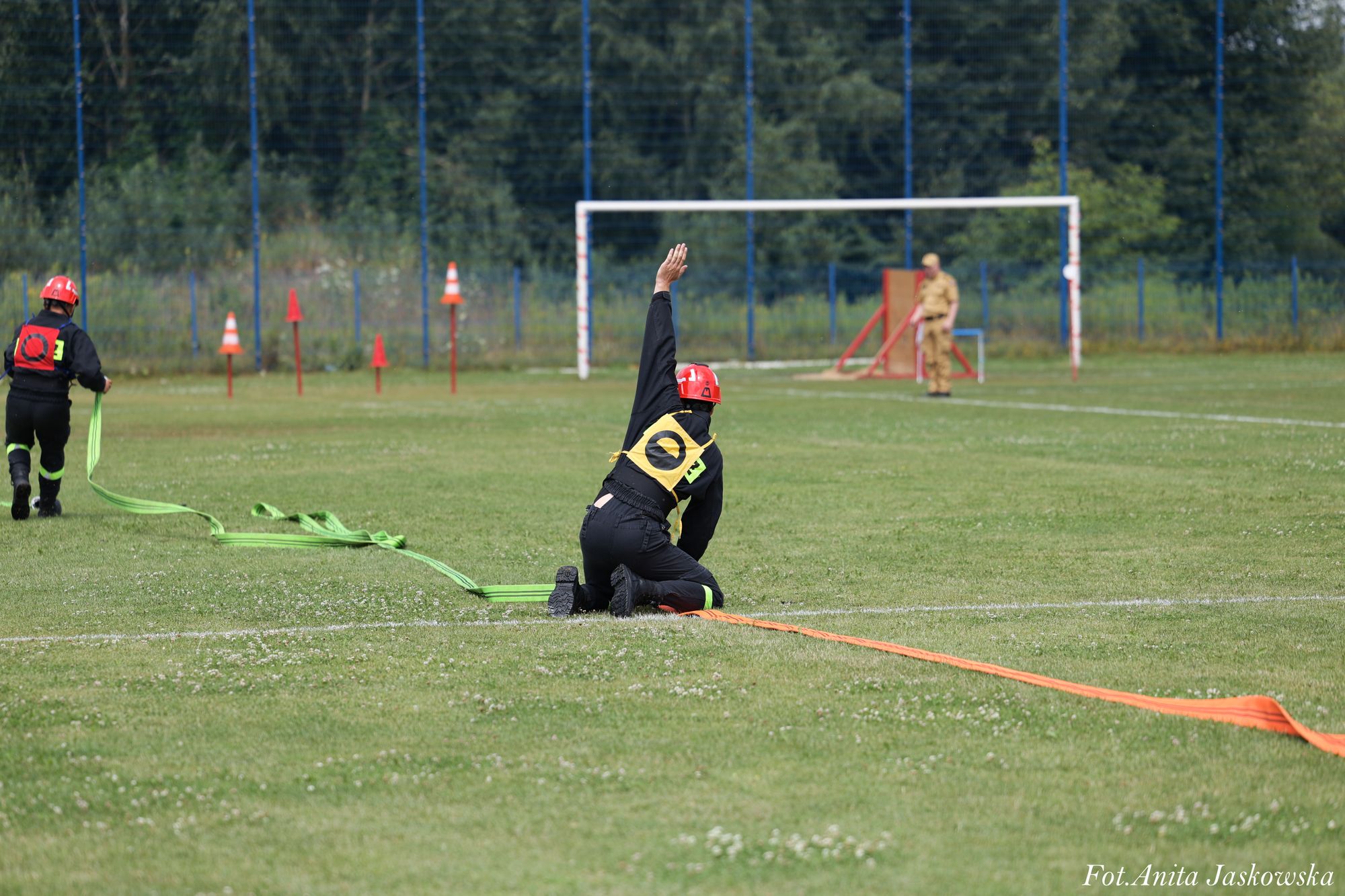 Dwie osoby w czarnych mundurach i czerwonych hełmach na zielonym boisku, jedna klęczy z podniesioną ręką, druga trzyma rozwinięty zielony wąż strażacki, w tle bramka i sylwetka osoby w mundurze beżowym.