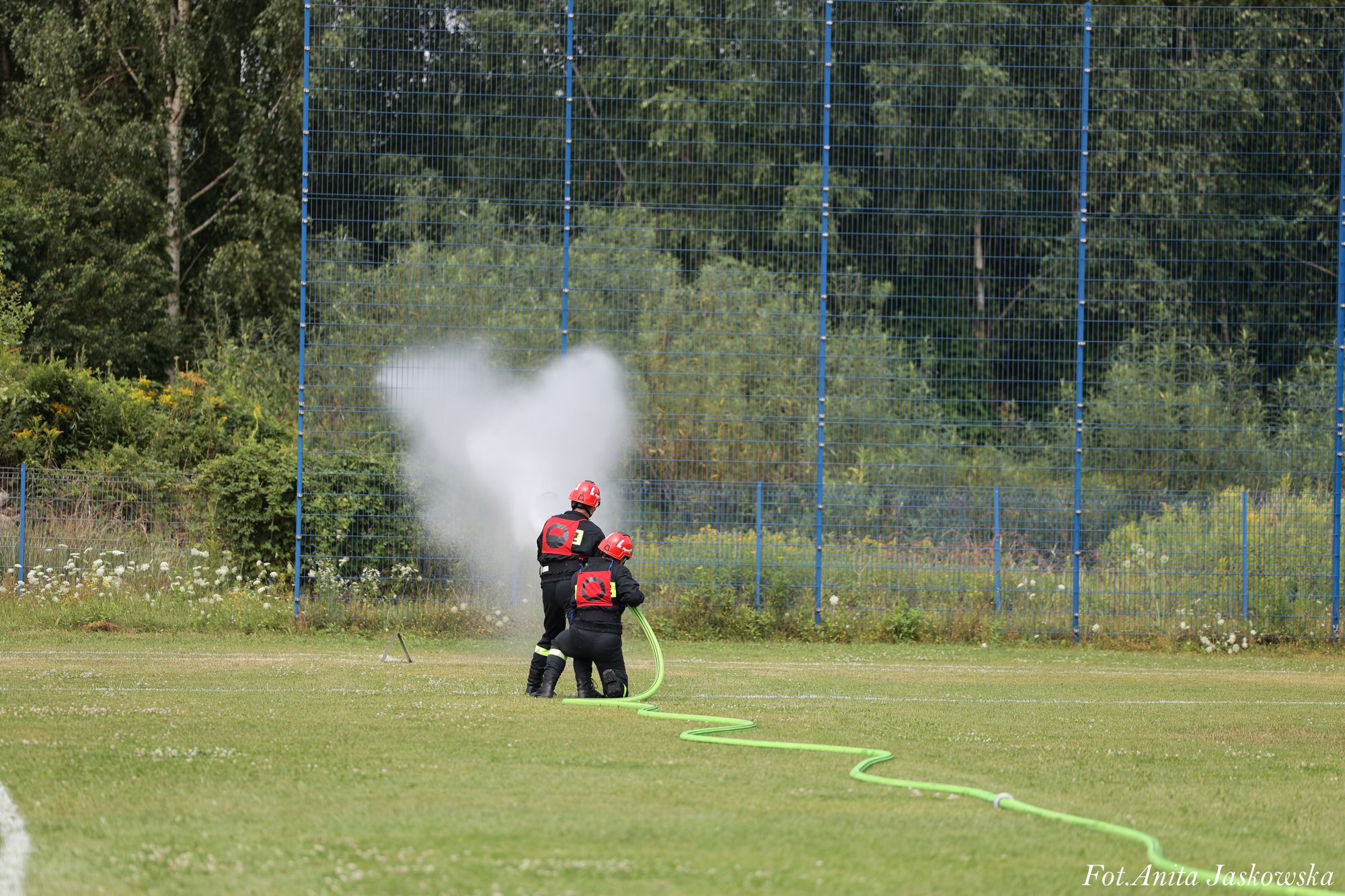 Dwóch strażaków w czarnych uniformach i czerwonych hełmach używa zielonego węża, z którego wypływa silny strumień wody na zielonym trawiastym boisku.