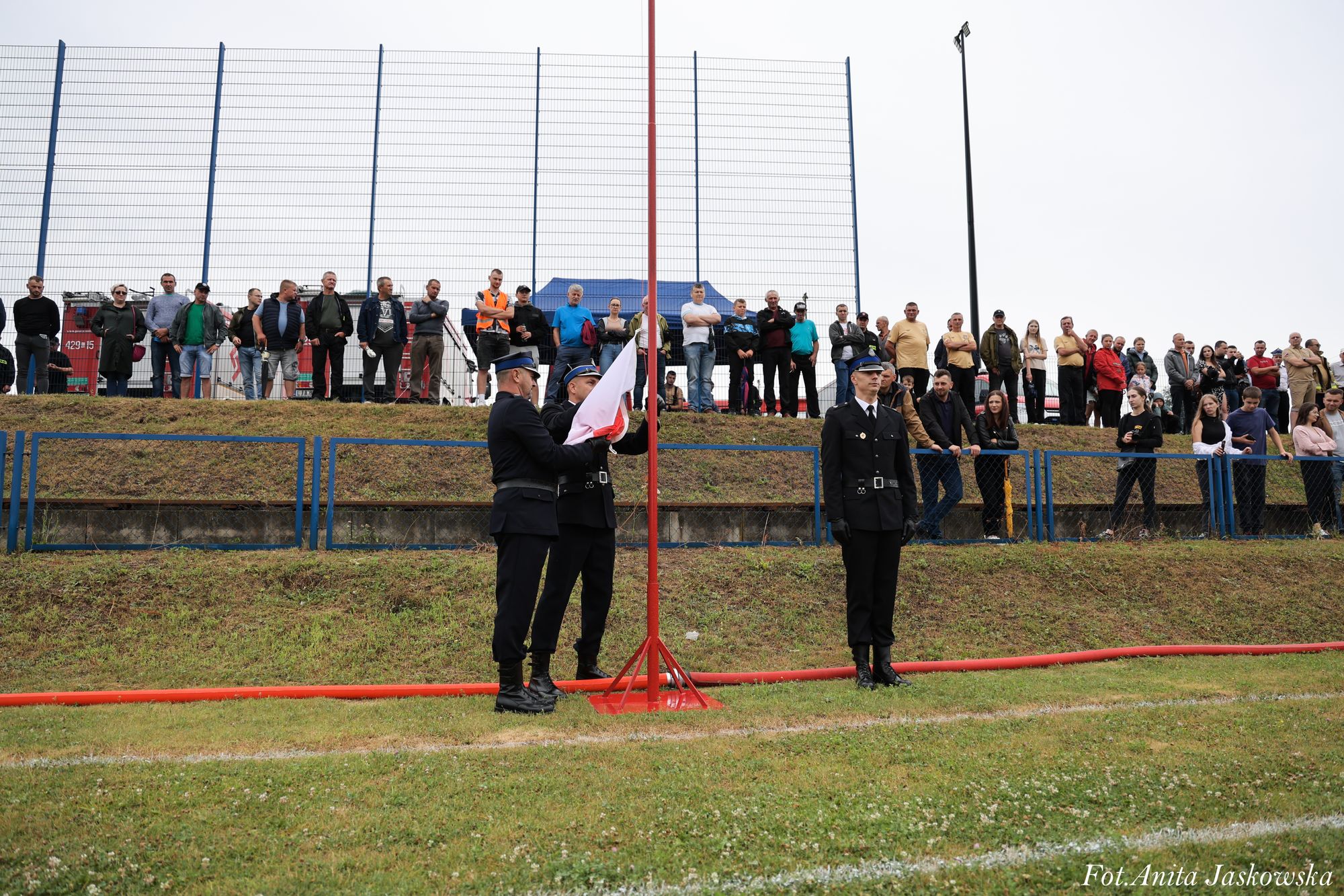 Trzech mężczyzn w mundurach podnosi flagę na maszcie na tle zielonej murawy, za nimi tłum ludzi stojących przy ogrodzeniu i metalowej siatce.