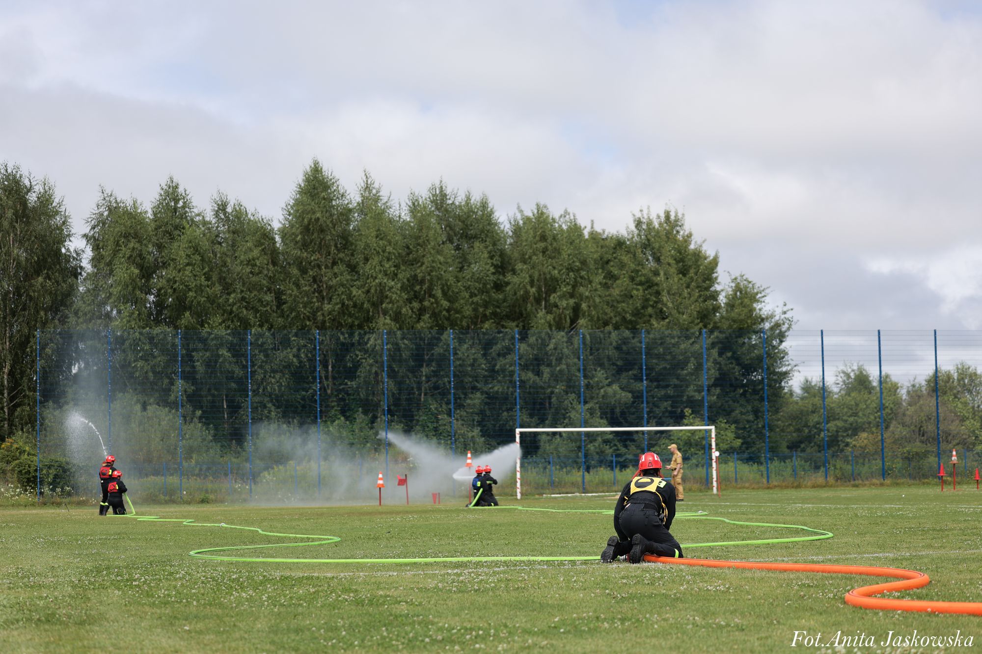 Trzech strażaków w czarnych uniformach i czerwonych hełmach na zielonym boisku, dwóch z nich używa węży do gaszenia, w tle bramka i drzewa.