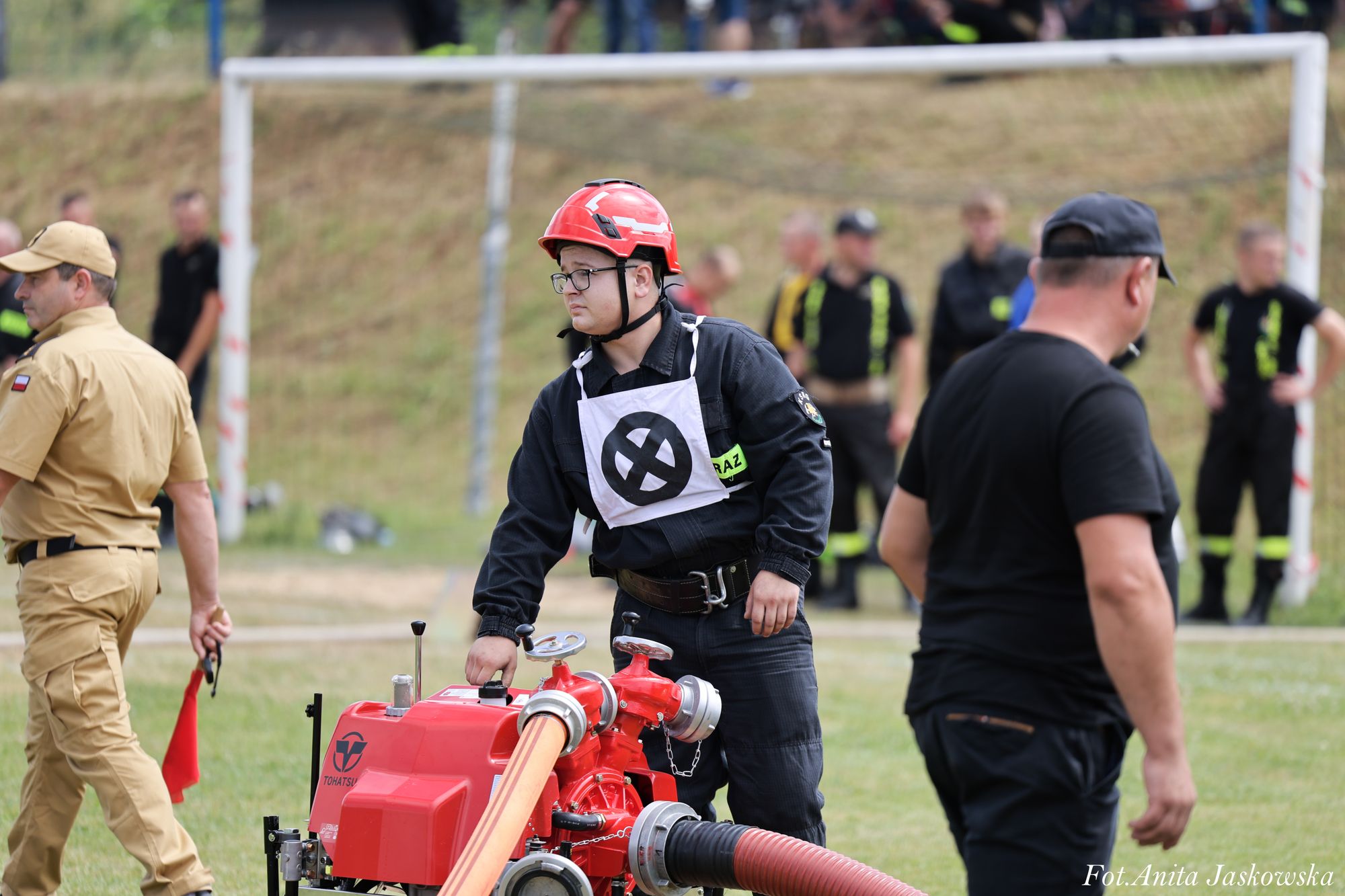 Mężczyzna w czarnym ubraniu i czerwonym hełmie obsługuje czerwony sprzęt z wężami na trawniku, w tle kilka osób i bramka do piłki nożnej.