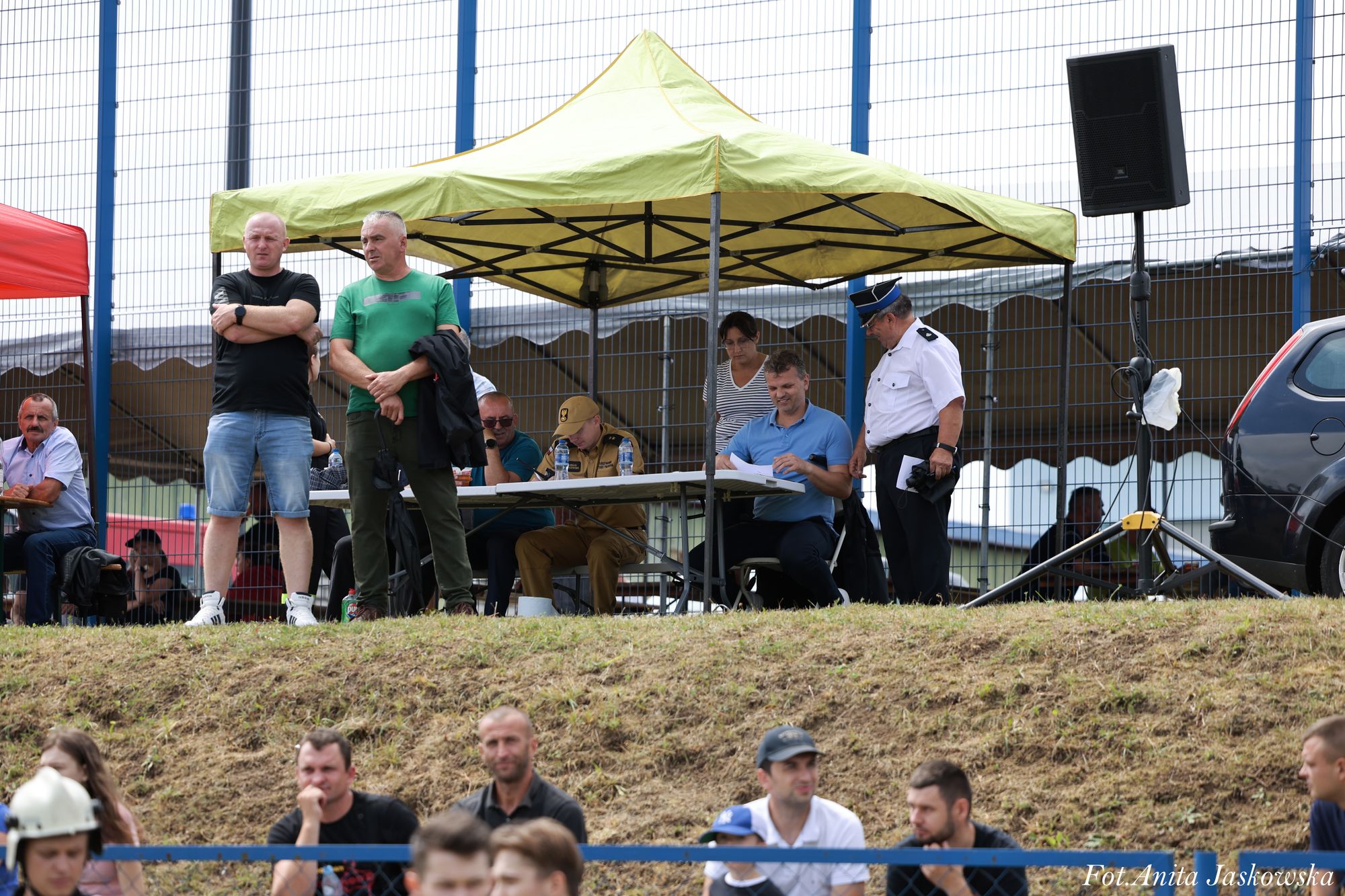 Grupa osób pod żółtym namiotem i wokół, z mężczyzną w mundurze oraz głośnikiem na stojaku obok niebieskiego ogrodzenia i samochodu.
