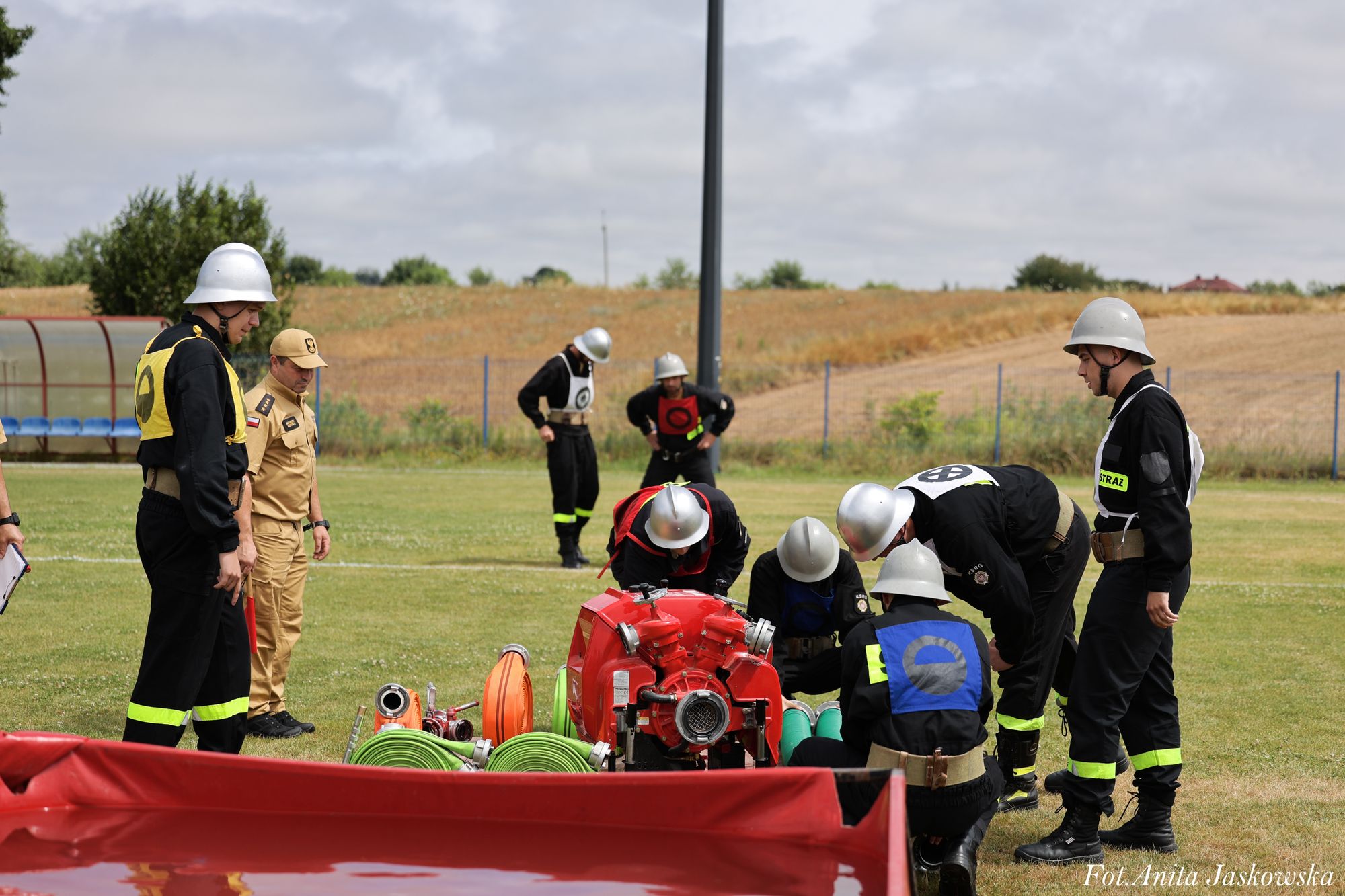 Grupa strażaków w czarnych mundurach i srebrnych hełmach wokół sprzętu pożarniczego na trawiastym polu, w tle sucha trawa i siatka ogrodzeniowa.