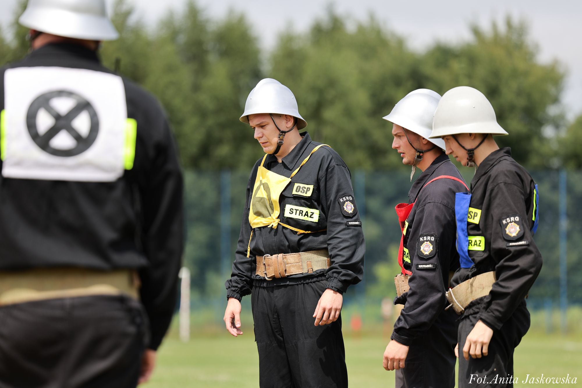 Czterech mężczyzn w czarnych mundurach strażackich z kolorowymi oznaczeniami, białe hełmy, stojący na zielonym polu na tle drzew.