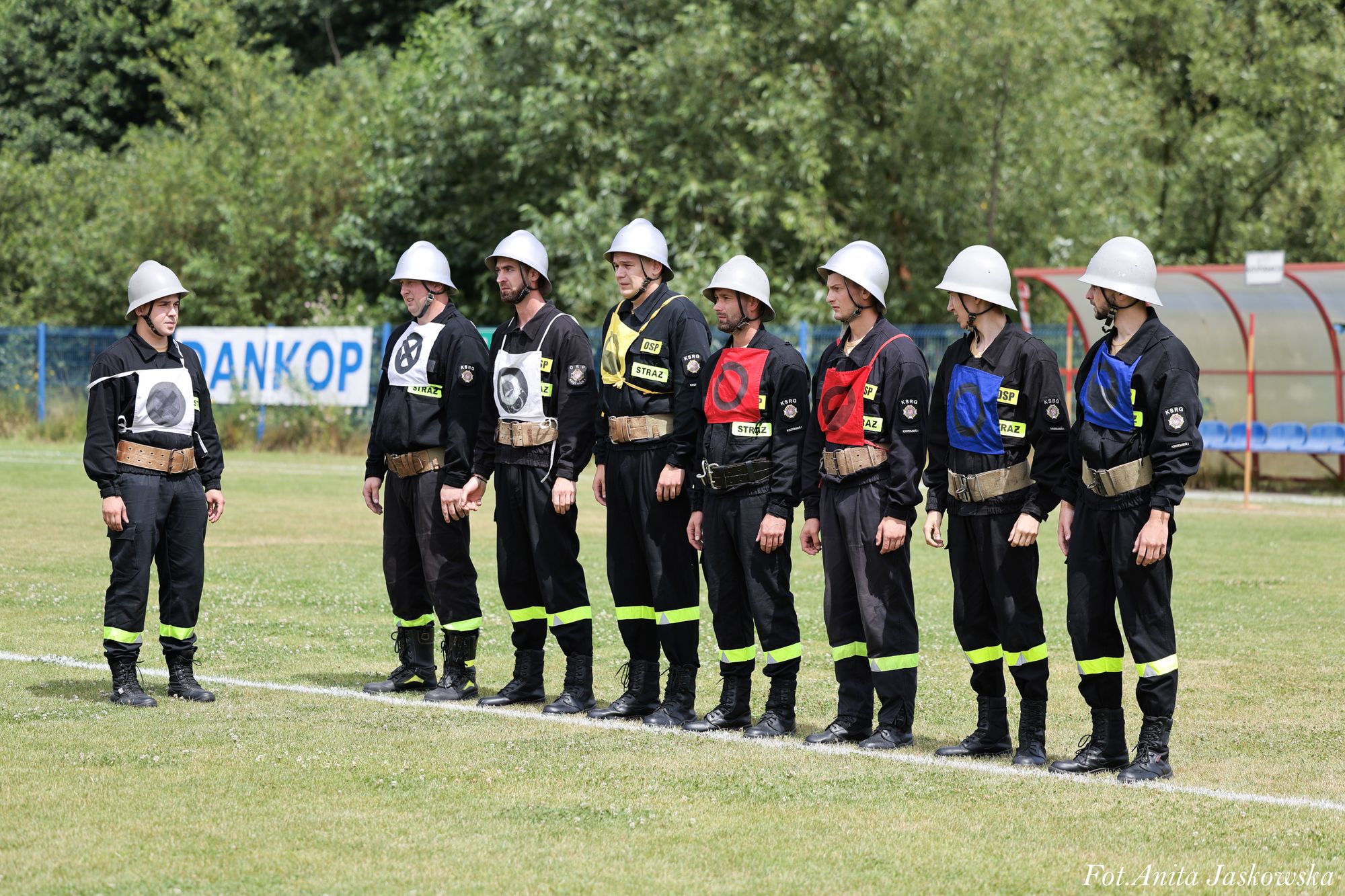 Grupa dziewięciu mężczyzn w czarnych uniformach z żółtymi odblaskowymi paskami, białych hełmach i kolorowych kamizelkach stojąca na trawiastym boisku, w tle drzewa i ławki.