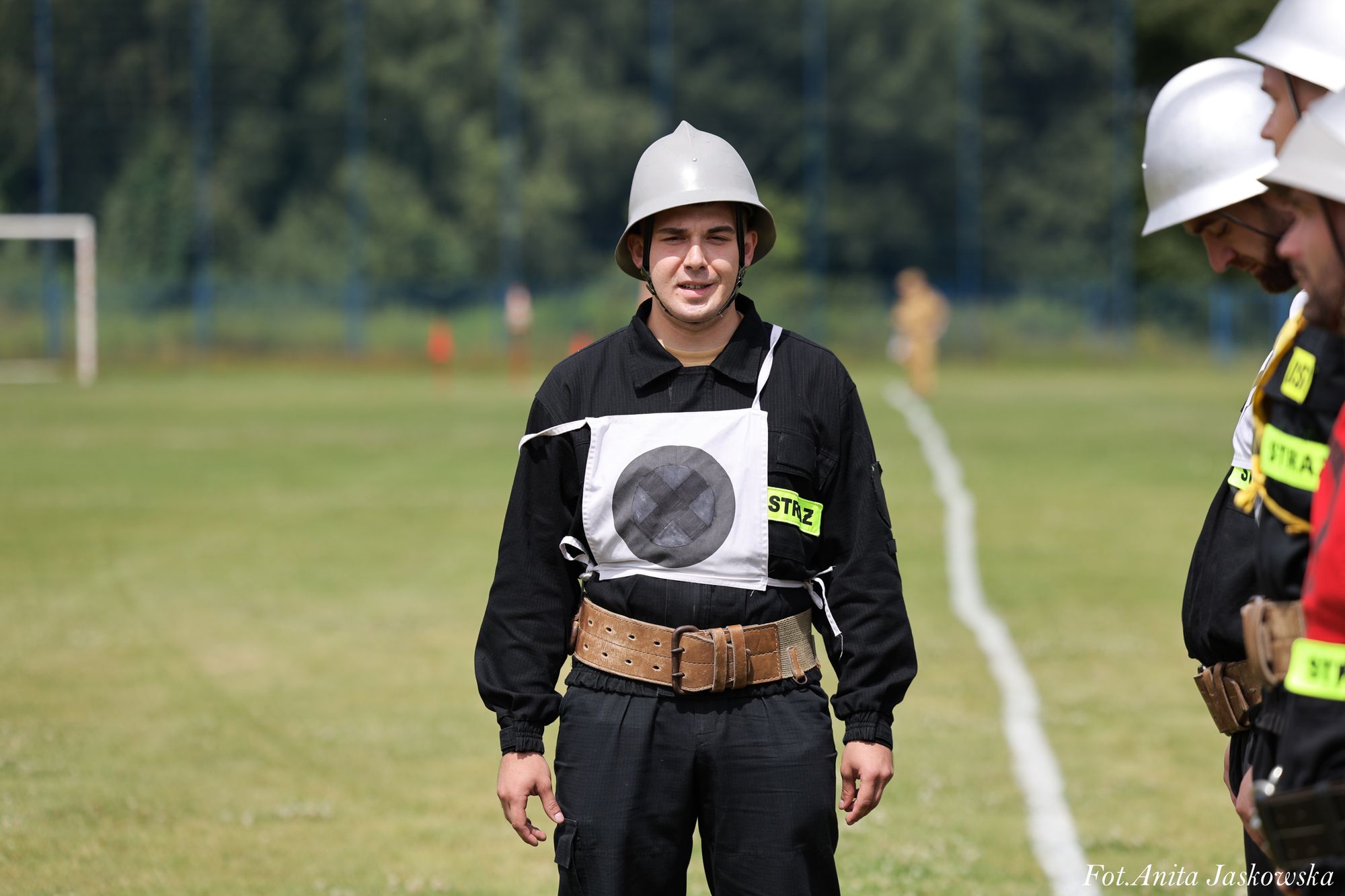 Mężczyzna w czarnym ubraniu strażackim, białym hełmie i białym oznaczeniu z czarnym kółkiem na piersi stoi na zielonym boisku, obok inni strażacy.