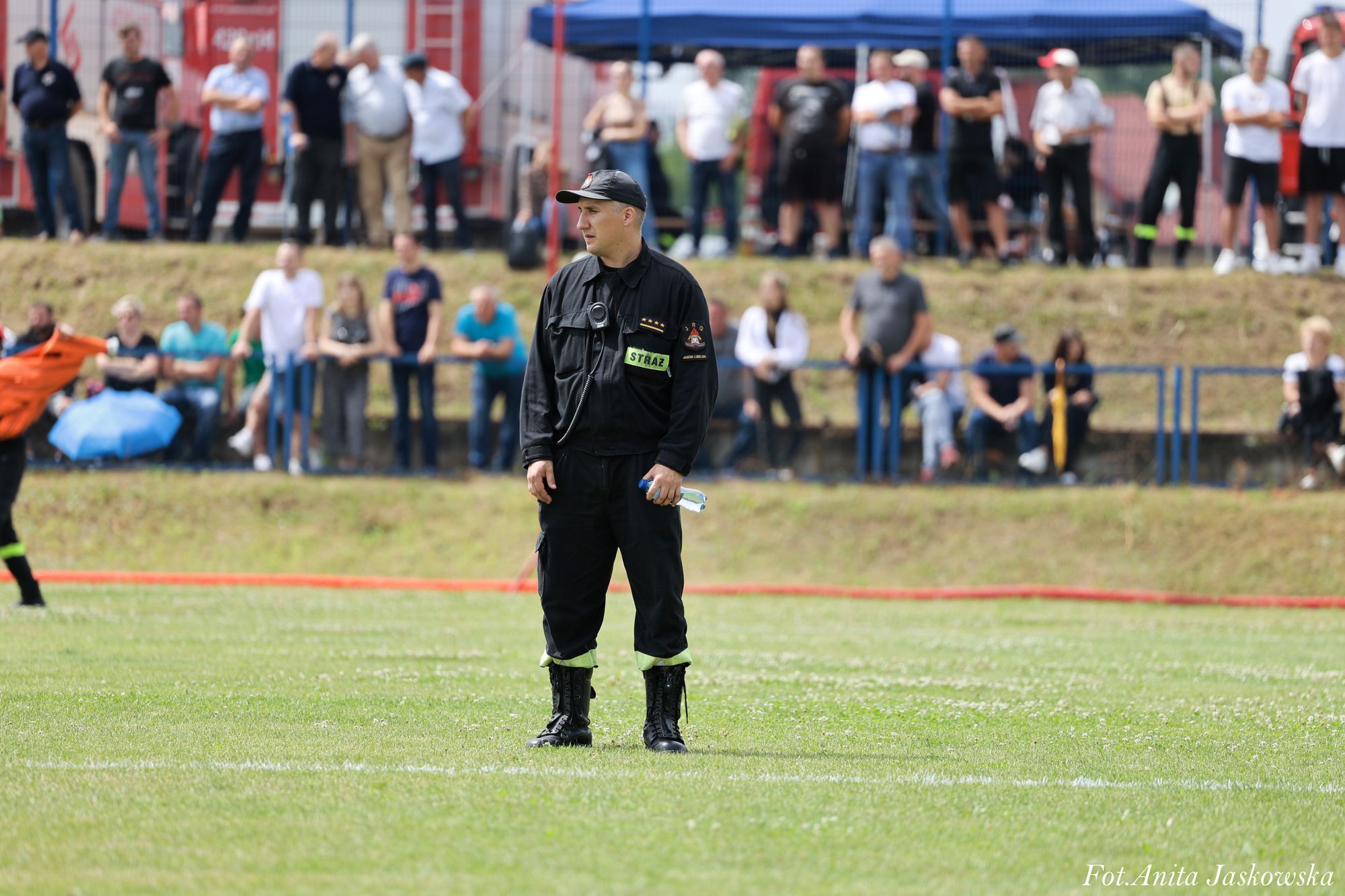 Mężczyzna w czarnym mundurze strażaka z napisem "STRAŻ" i czapką stoi na zielonym boisku, w tle rozmyci ludzie na trybunach i niebieski namiot.