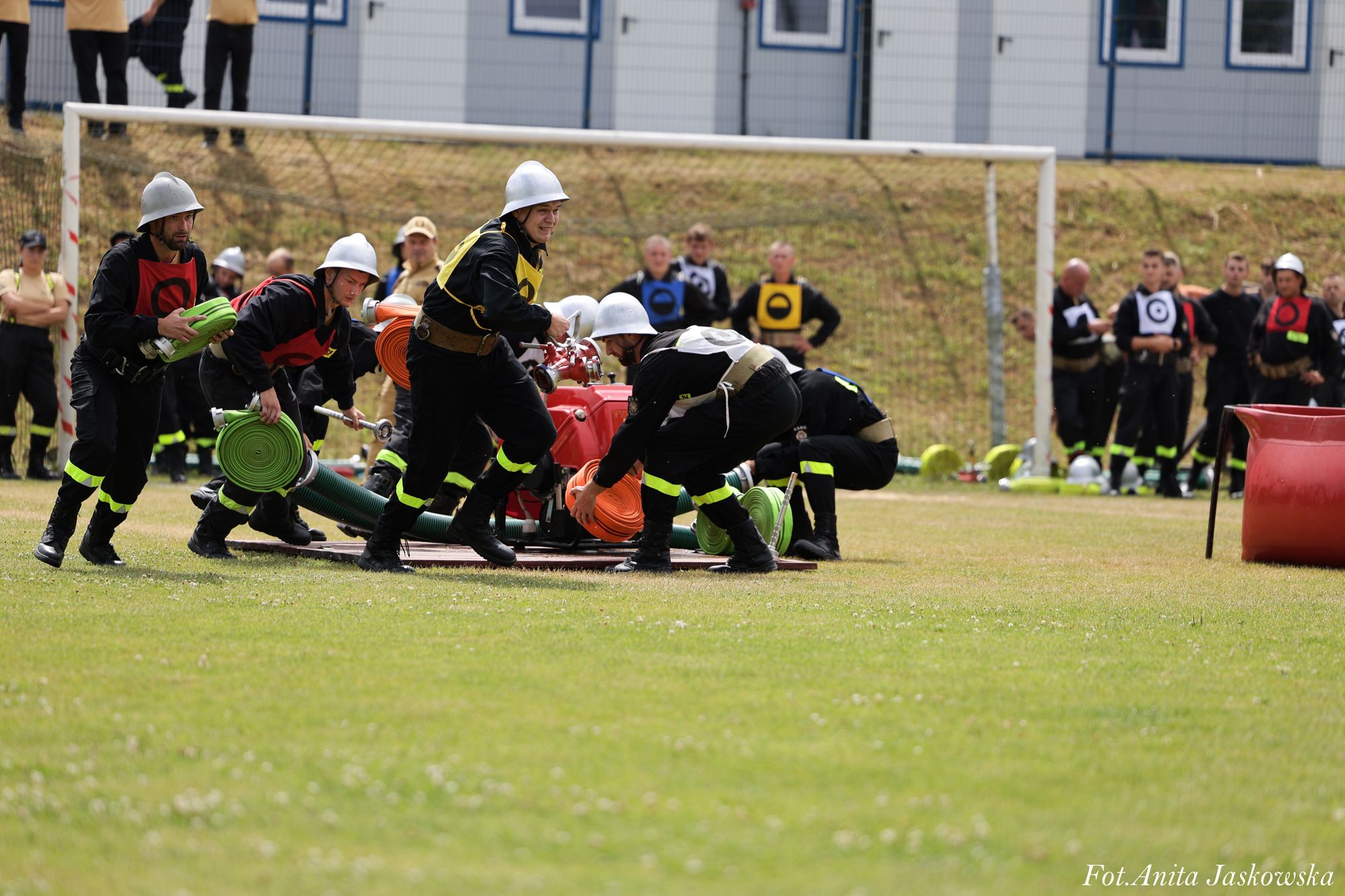 Grupa strażaków w hełmach i ubraniach ochronnych rozwija zielone i pomarańczowe węże położone na trawie na tle bramki i widowni.