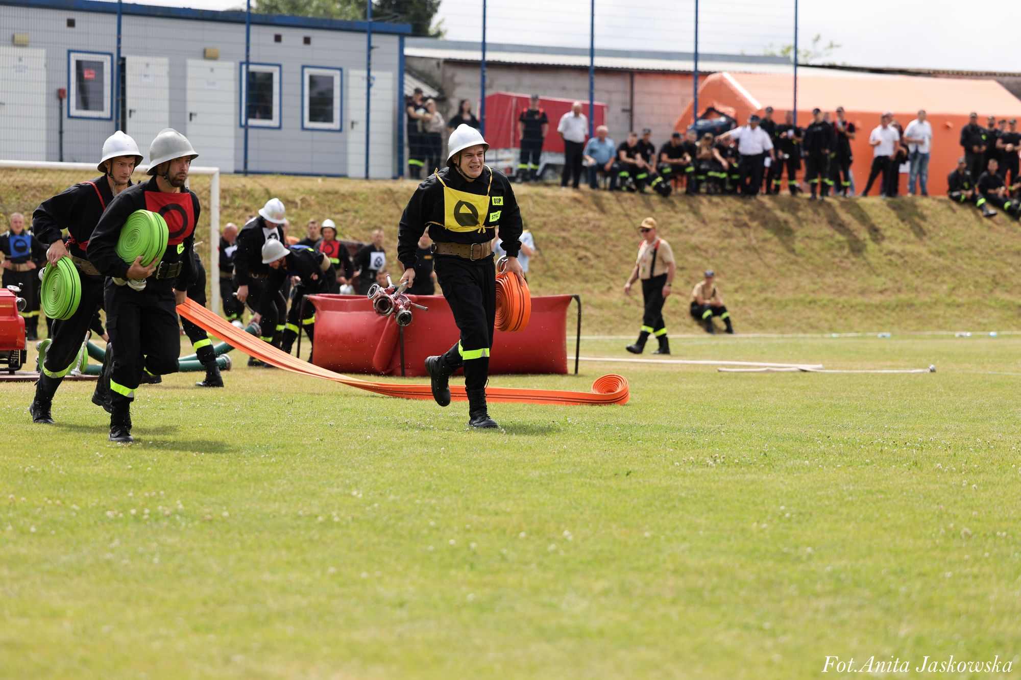 Strażacy w czarnych mundurach i białych hełmach biegną z zielonymi i pomarańczowymi zwiniętymi wężami na zielonej trawie, w tle widoczna grupa ludzi i kontenery.