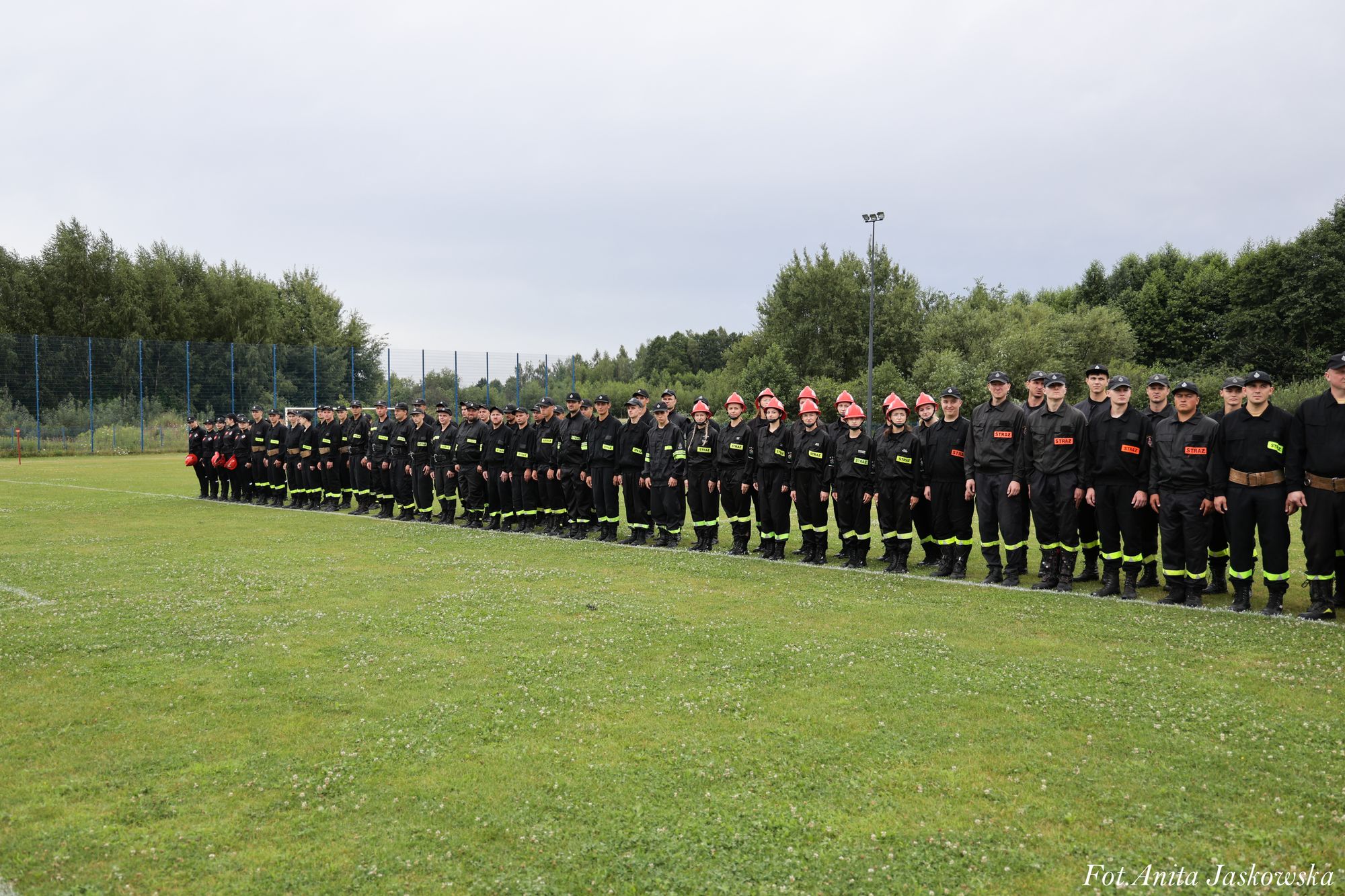 Grupa osób w czarnych mundurach z żółtymi odblaskami stoi w jednej linii na zielonej trawie, w tle drzewa i ogrodzenie boiska.
