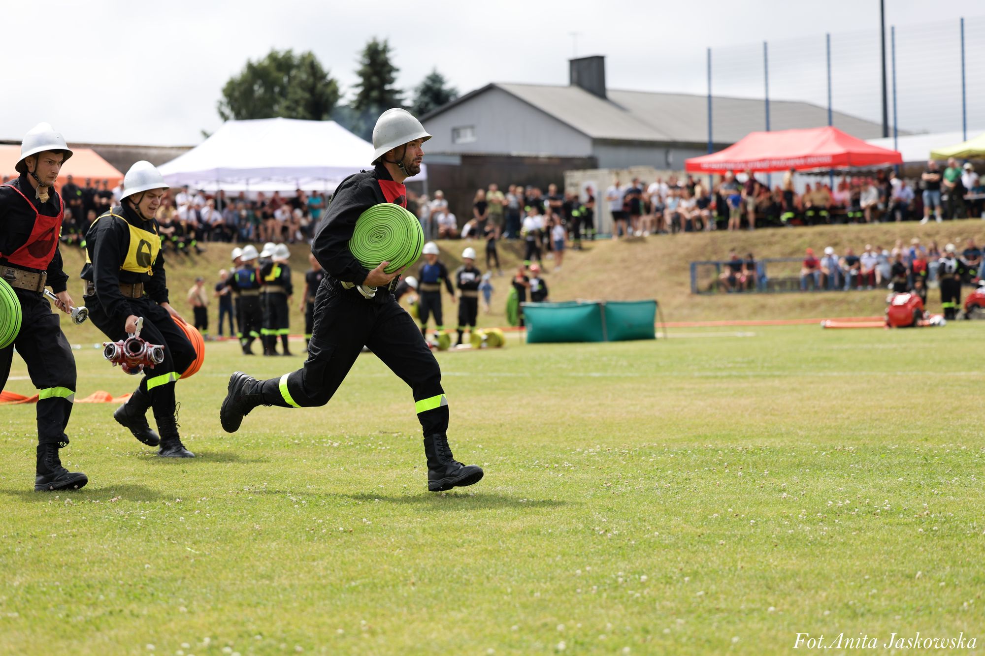 Trzech mężczyzn w czarnych ubraniach strażackich i hełmach biegnie po zielonej trawie, trzymając zwinięte zielone i pomarańczowe węże strażackie, w tle widzowie i namioty.