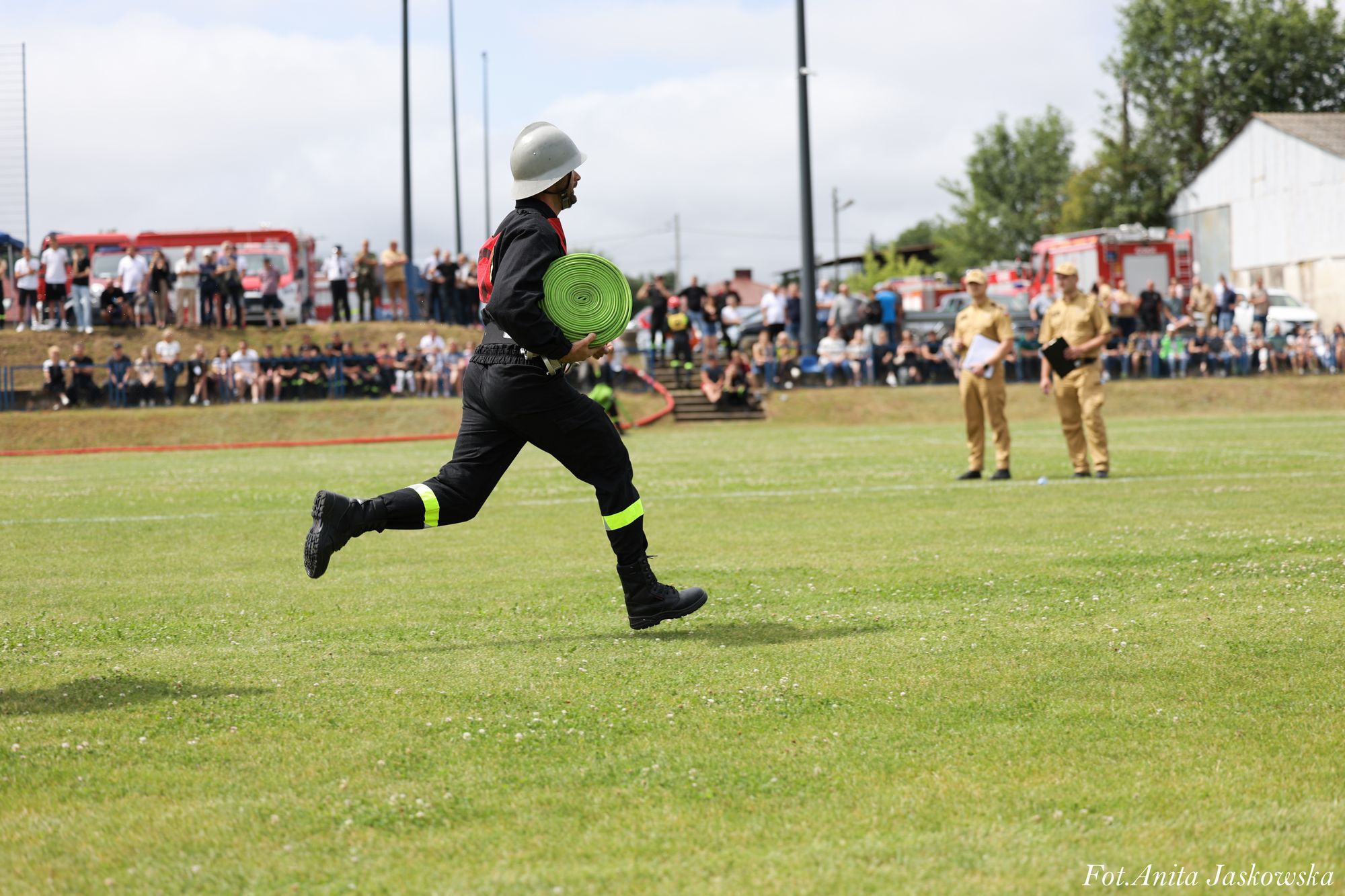 Strażak w czarnym ubraniu z odblaskowymi paskami i białym hełmem biegnie po zielonej trawie, trzymając zielony zwinięty wąż strażacki, w tle tłum ludzi i wozy strażackie.