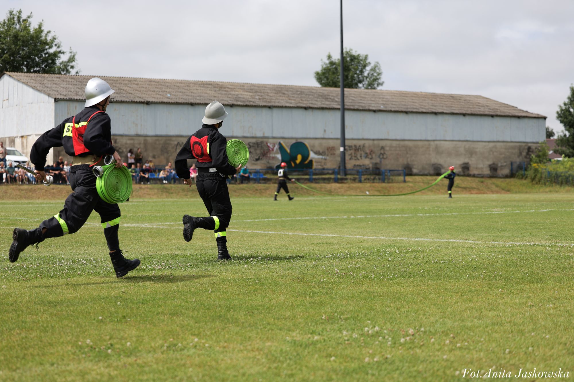 Czterej strażacy w czarnych mundurach i hełmach biegną po zielonym boisku, dwóch trzyma zwinięte zielone węże strażackie, w tle budynek i widzowie.