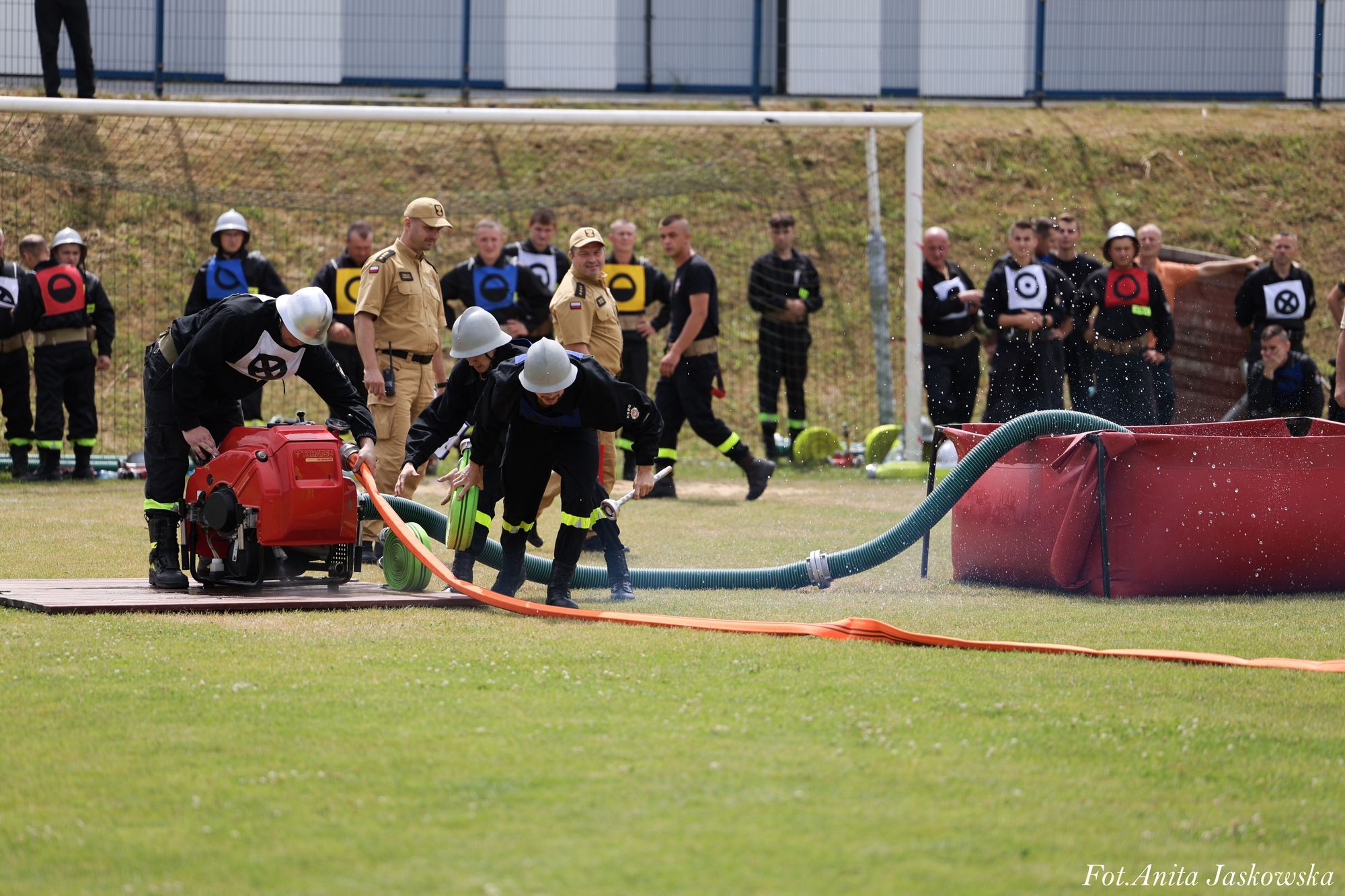 Strażacy w czarnych mundurach i srebrnych hełmach rozkładają wąż połączony z czerwoną maszyną i zbiornikiem na trawiastym boisku, w tle ludzie w mundurach obserwują ćwiczenia.