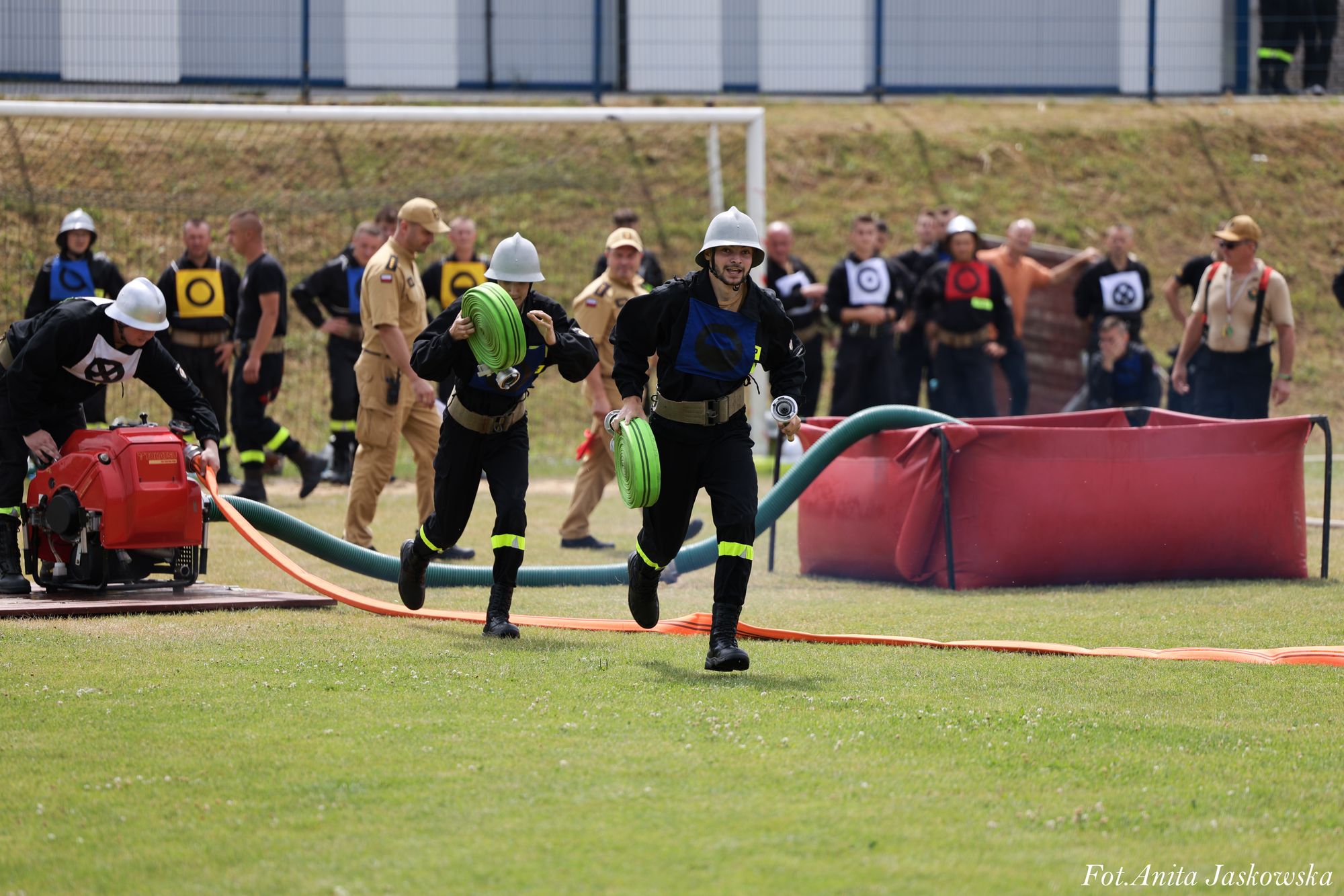 Grupa strażaków w hełmach, dwóch biegnących z zielonymi zwiniętymi wężami, czerwony zbiornik i pompa, na tle trawy i ogrodzenia.