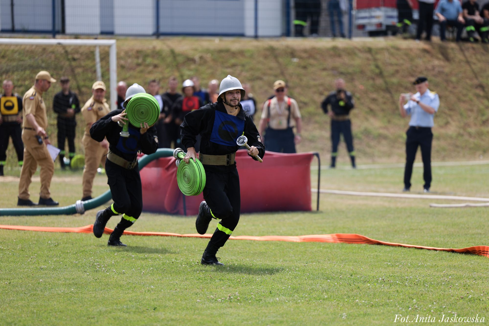 Dwóch strażaków w kaskach i czarnych uniformach biegnie po zielonej trawie, niosąc zielone zwinięte węże przeciwpożarowe, w tle ludzie obserwujący zawody.