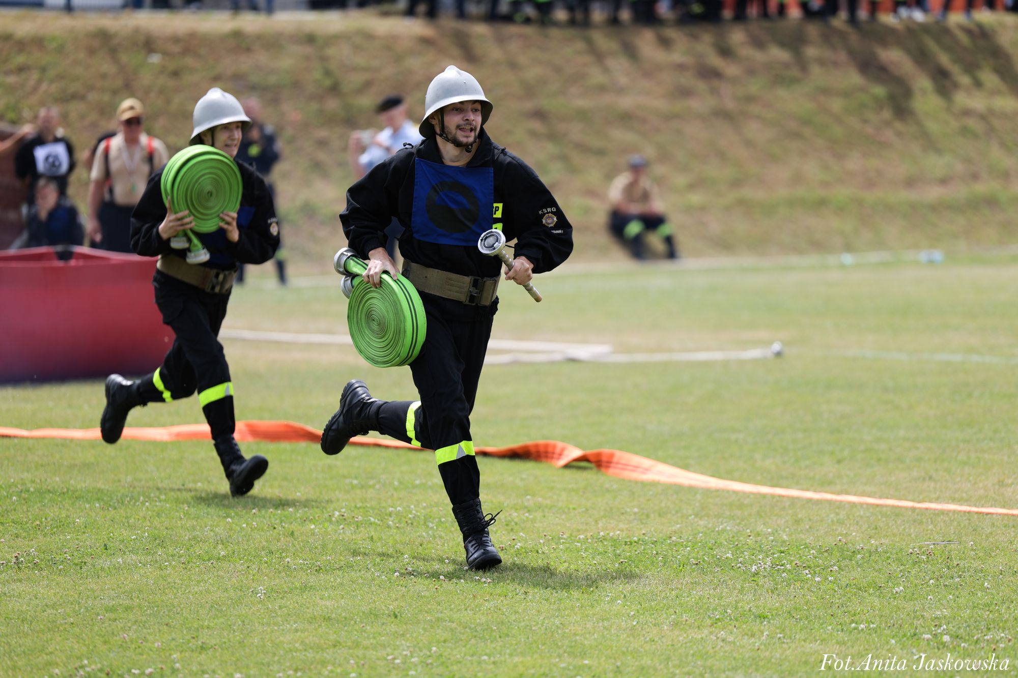 Dwóch mężczyzn w czarnych mundurach i białych hełmach biegnie po zielonej trawie, każdy niesie zielony zwinięty wąż strażacki. Tło rozmyte.