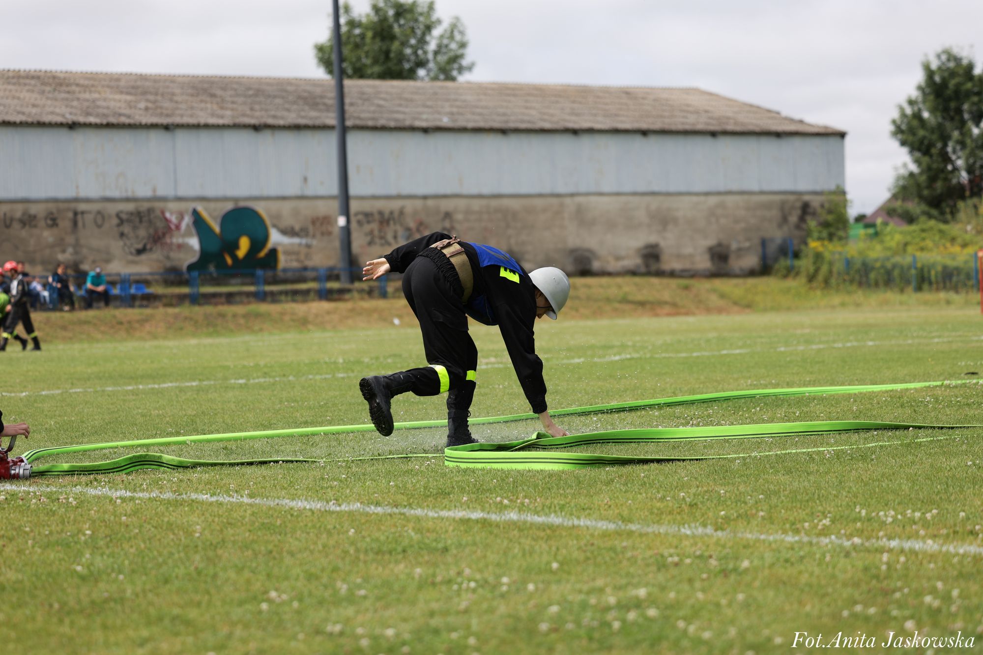 Strażak w czarnym ubraniu z odblaskowymi paskami i białym hełmem rozkłada zielony wąż strażacki na trawiastym boisku.