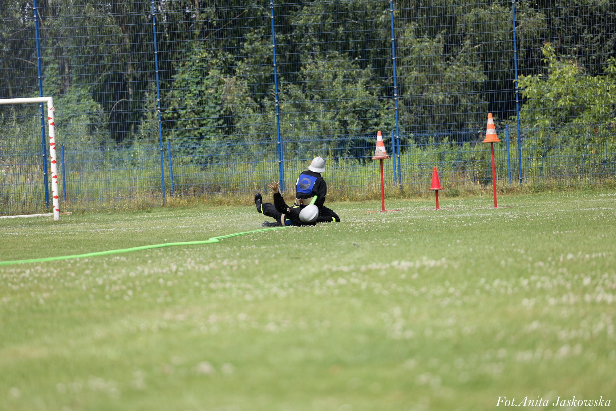 Dwóch ludzi w kaskach i ciemnych ubraniach na trawiastej murawie, zielony wąż, metalowy płot i pomarańczowe pachołki na tle drzew.