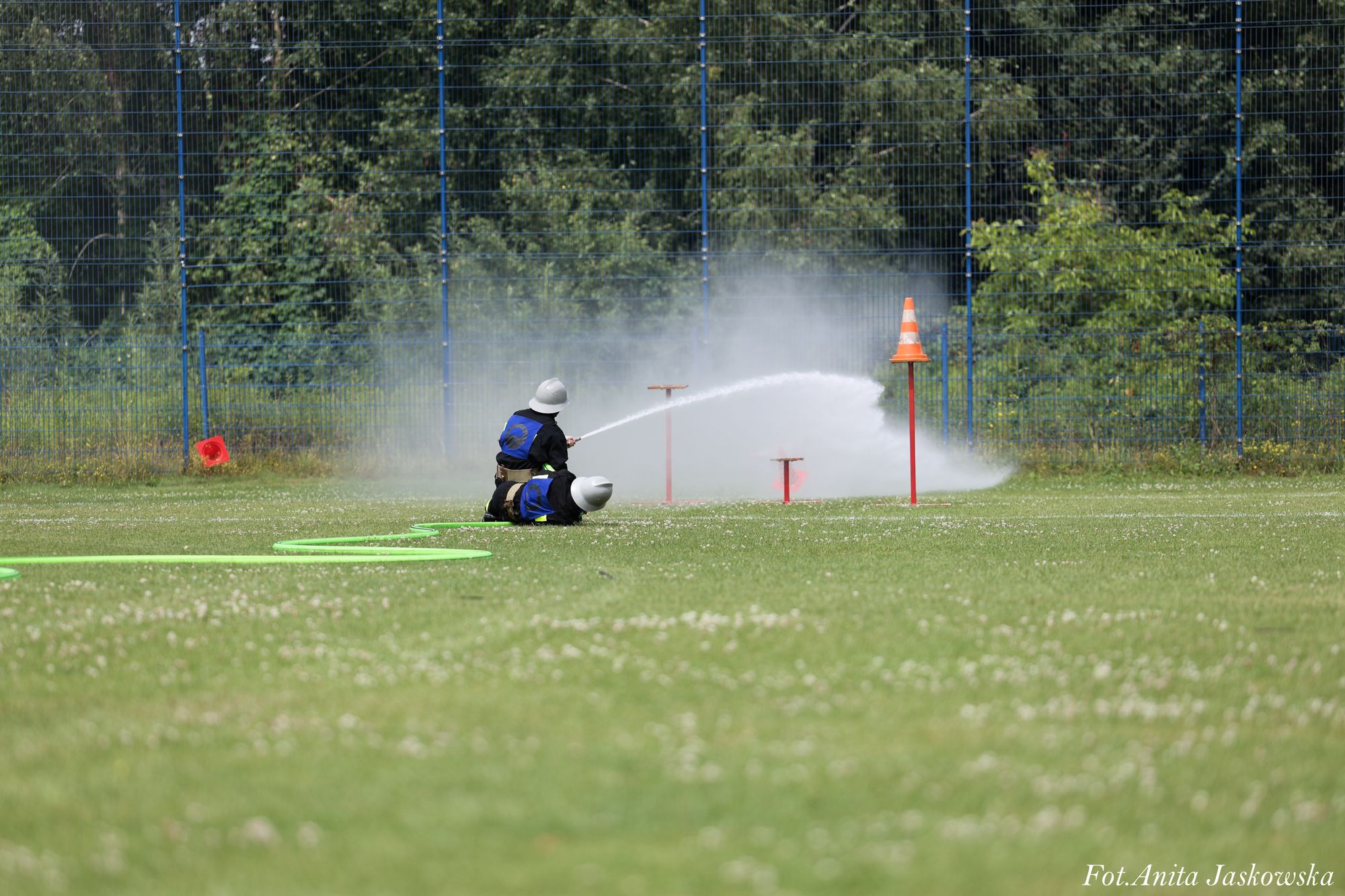 Dwoje strażaków w hełmach i niebieskich mundurach na zielonym trawniku, jeden z nich trzyma strumień wody skierowany na pachołek, za nimi niebieski metalowy płot i zieleń.