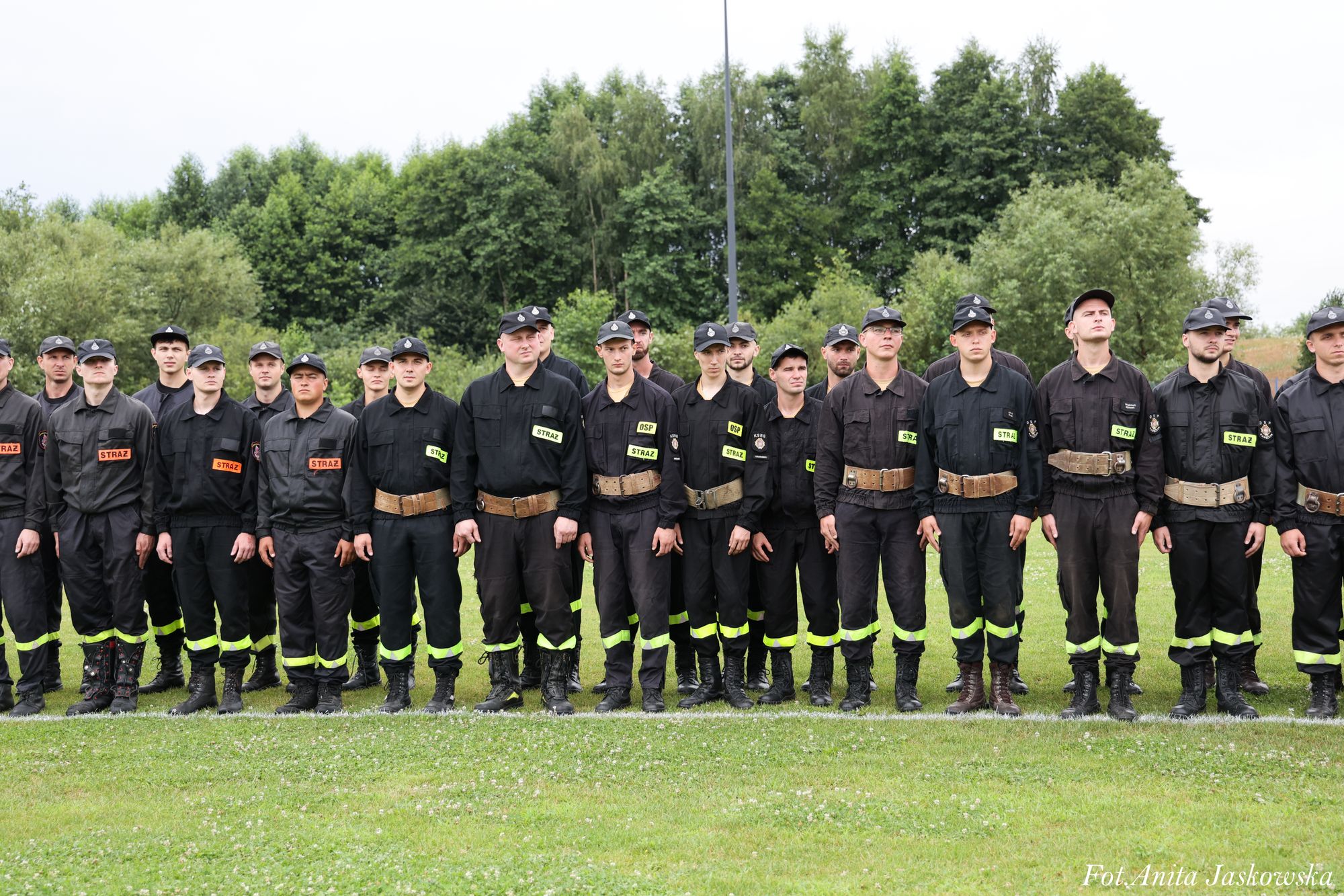 Grupa mężczyzn ubranych w ciemne mundury z odblaskowymi pasami i napisami "STRAŻ", stojących na zielonej trawie, za nimi drzewa i jasne niebo.
