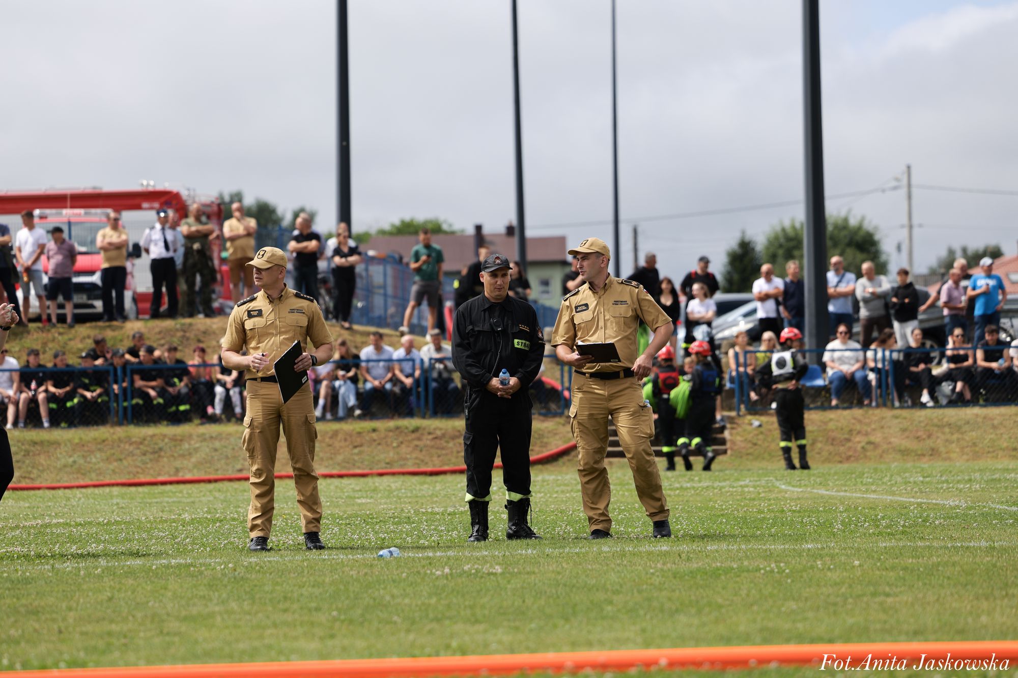 Trzech mężczyzn w mundurach, dwóch w beżowych, jeden w czarnym, stoi na trawiastym boisku, w tle grupa ludzi za ogrodzeniem i czerwony wóz strażacki.
