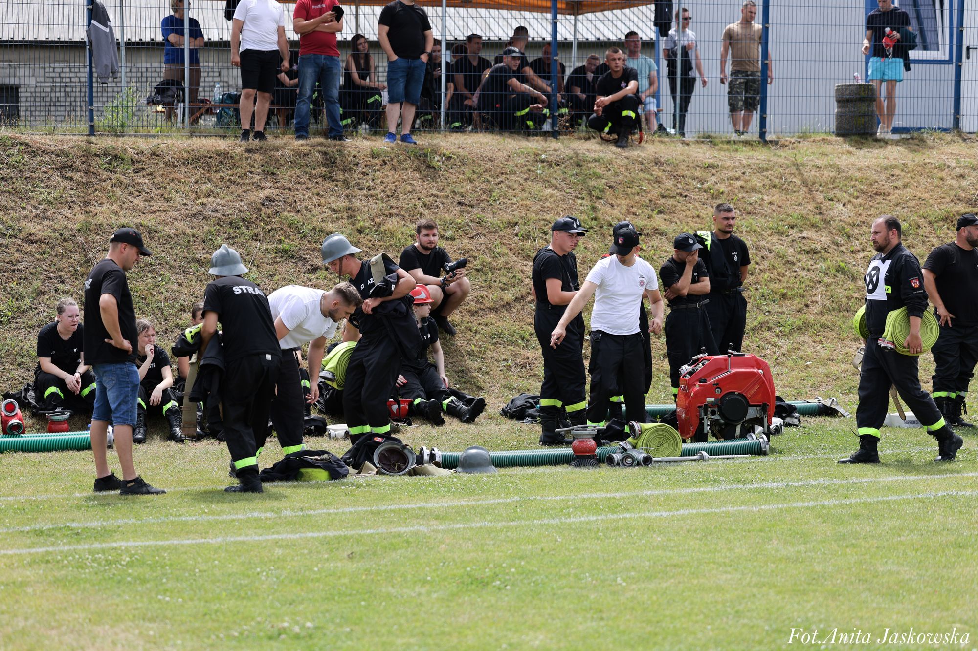 Grupa osób, głównie mężczyzn, ubranych w czarne ubrania strażackie z żółtymi odblaskami, stojących i siedzących na trawie z wężami strażackimi i sprzętem obok nich, w tle metalowe ogrodzenie i ludzie.