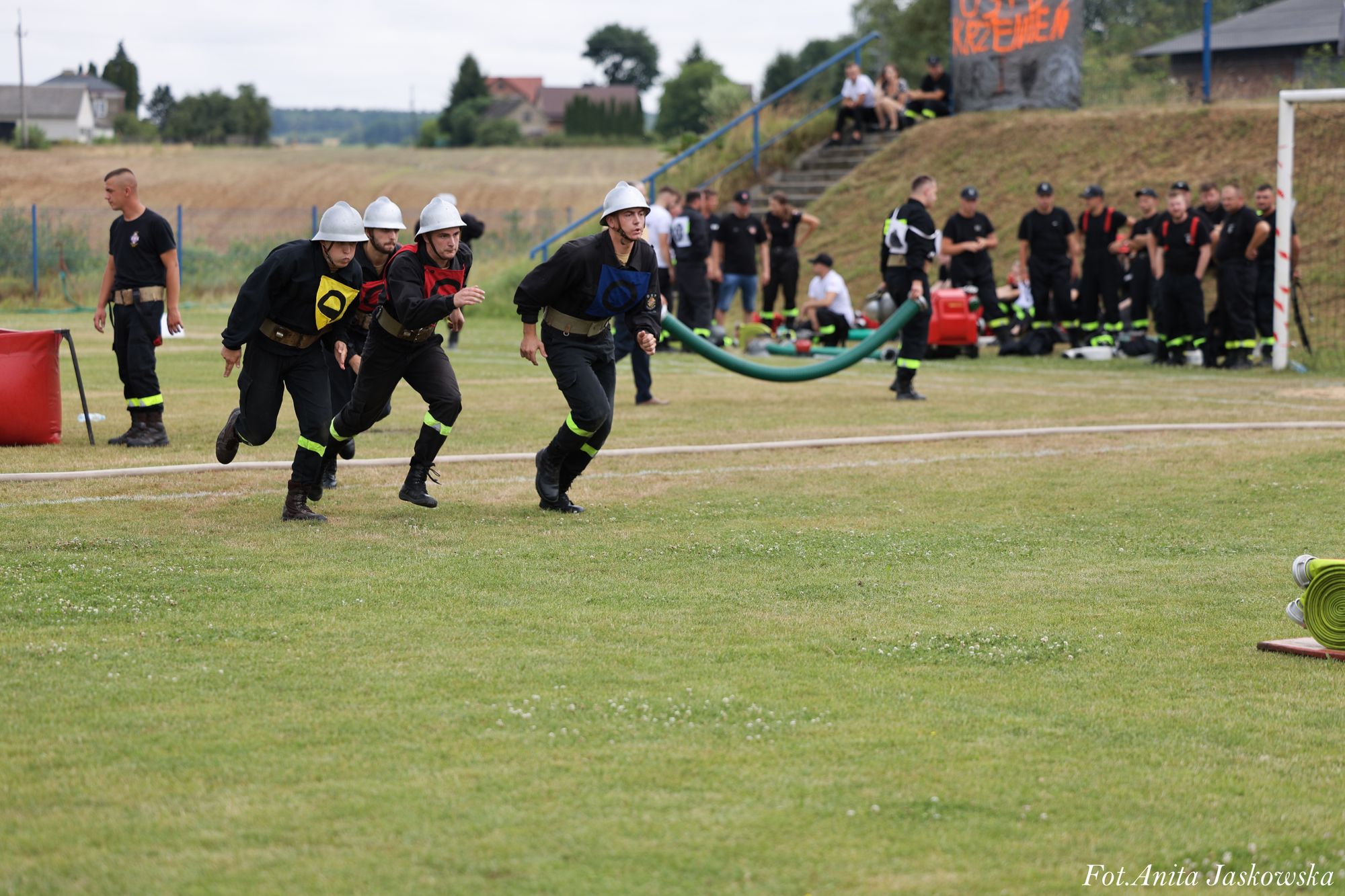 Czterech strażaków w hełmach i mundurach z kolorowymi oznaczeniami biegnie po trawiastym boisku, w tle grupa osób w mundurach i sprzęt strażacki.