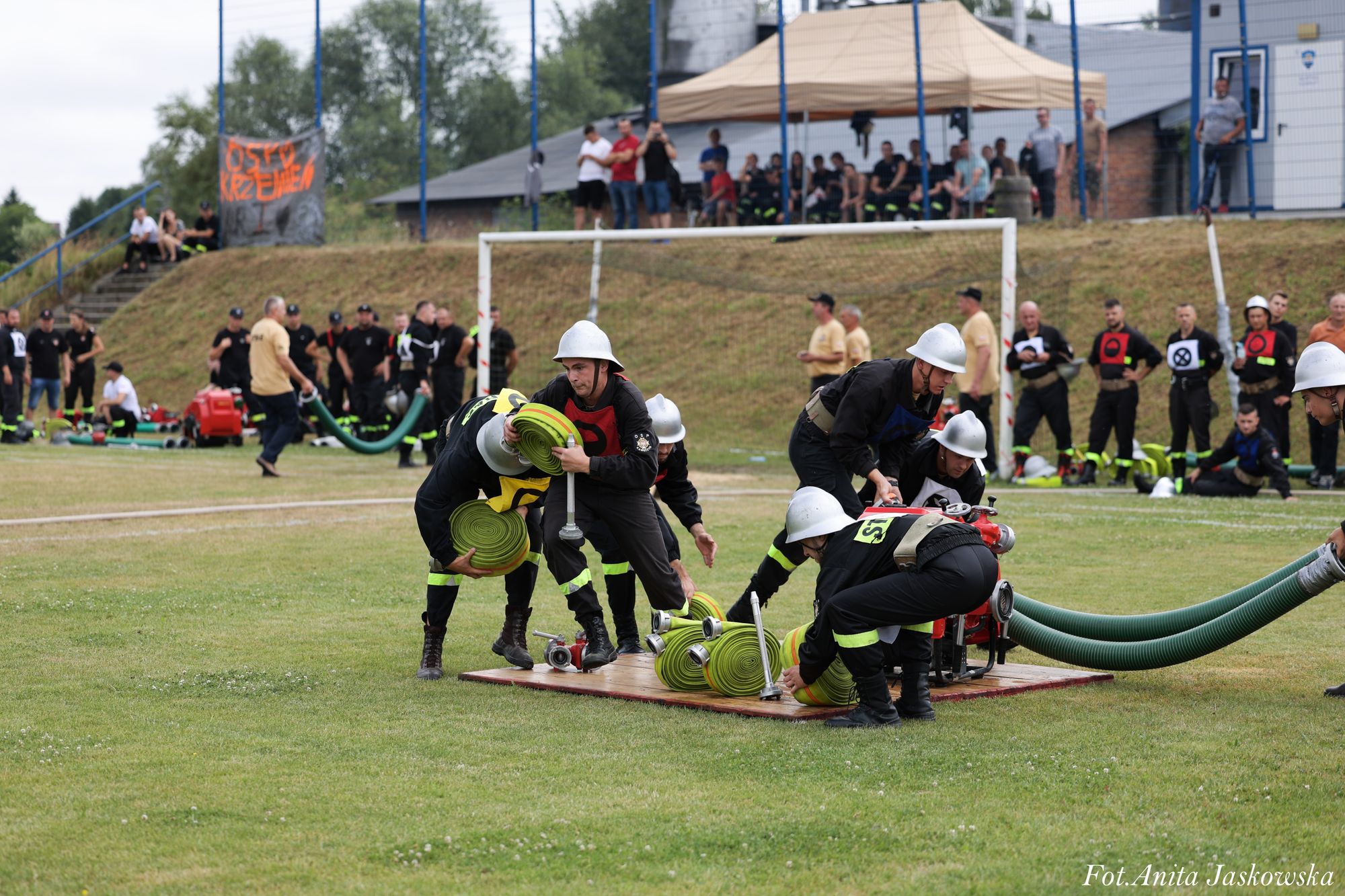 Grupa strażaków w hełmach i czarnych mundurach z żółtymi pasami rozwija zielone zwinięte węże na trawie przed bramką na boisku sportowym.