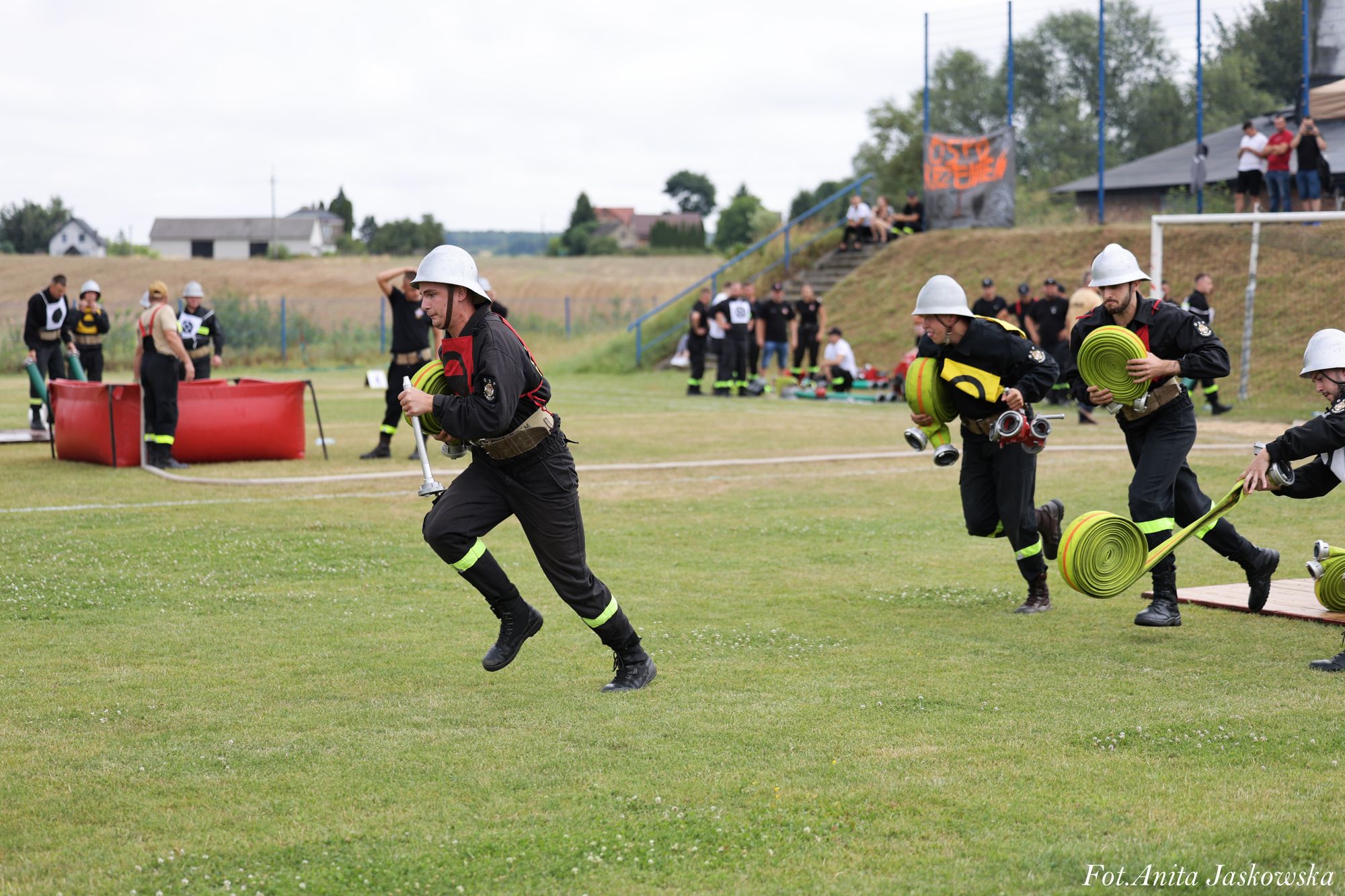 Strażacy w czarnych mundurach z żółtymi odblaskami i białych hełmach biegną po zielonym boisku trzymając zwinięte zielone węże strażackie, w tle ludzie i czerwony zbiornik.