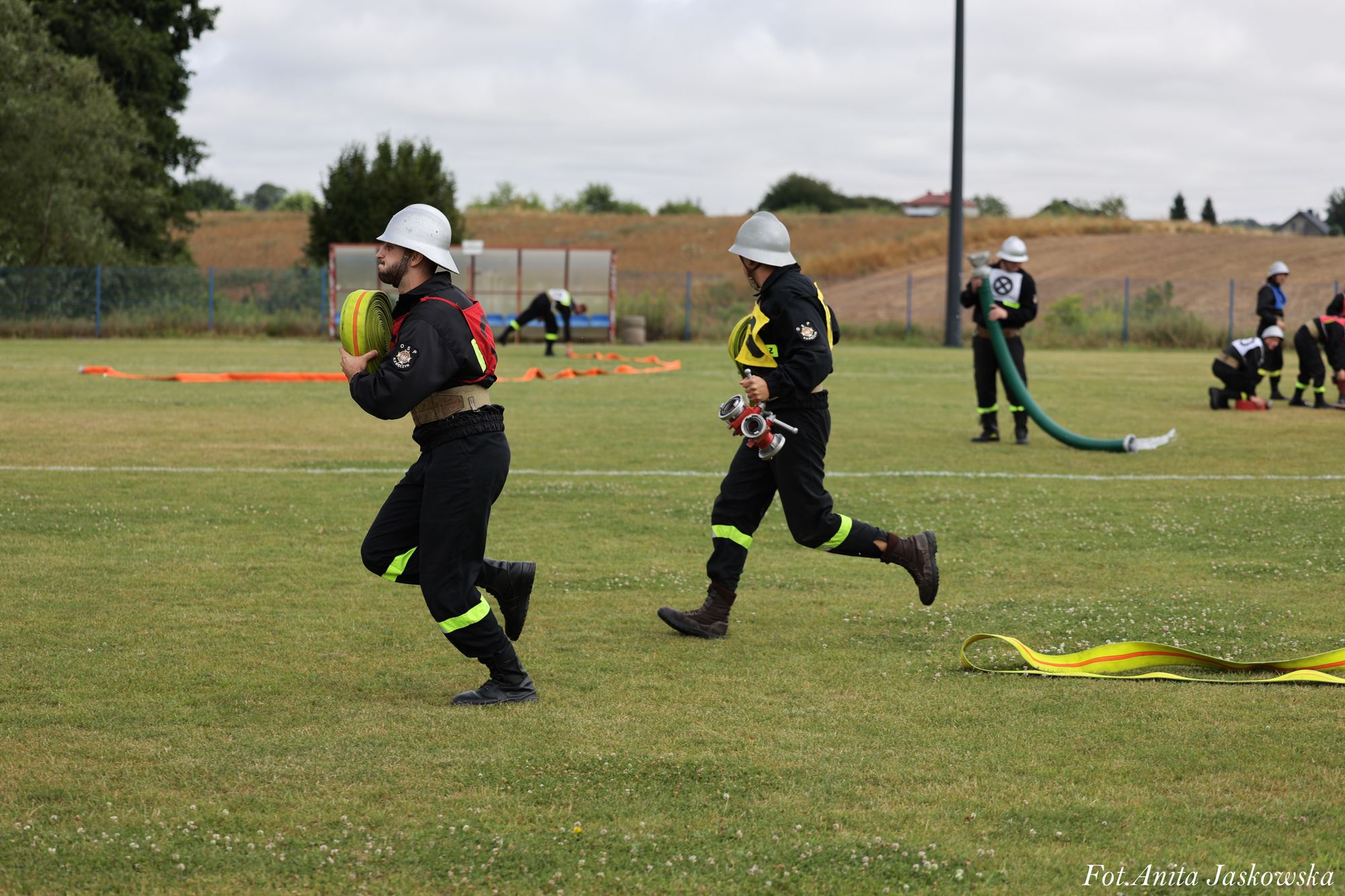 Dwóch strażaków w czarnych mundurach i białych hełmach biegnie po zielonej trawie, trzymając zwinięte węże strażackie, w tle inni strażacy i pola uprawne.