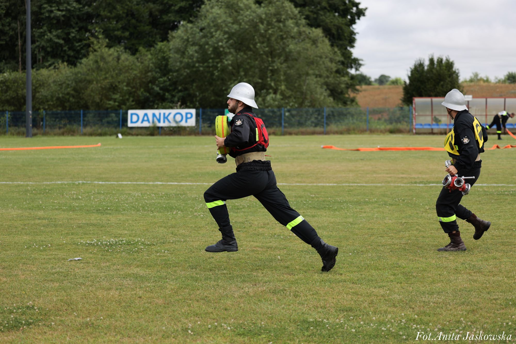Dwóch strażaków w czarnych mundurach z odblaskowymi paskami i białych hełmach biegnie po zielonym polu trawiastym, trzymając sprzęt pożarniczy.