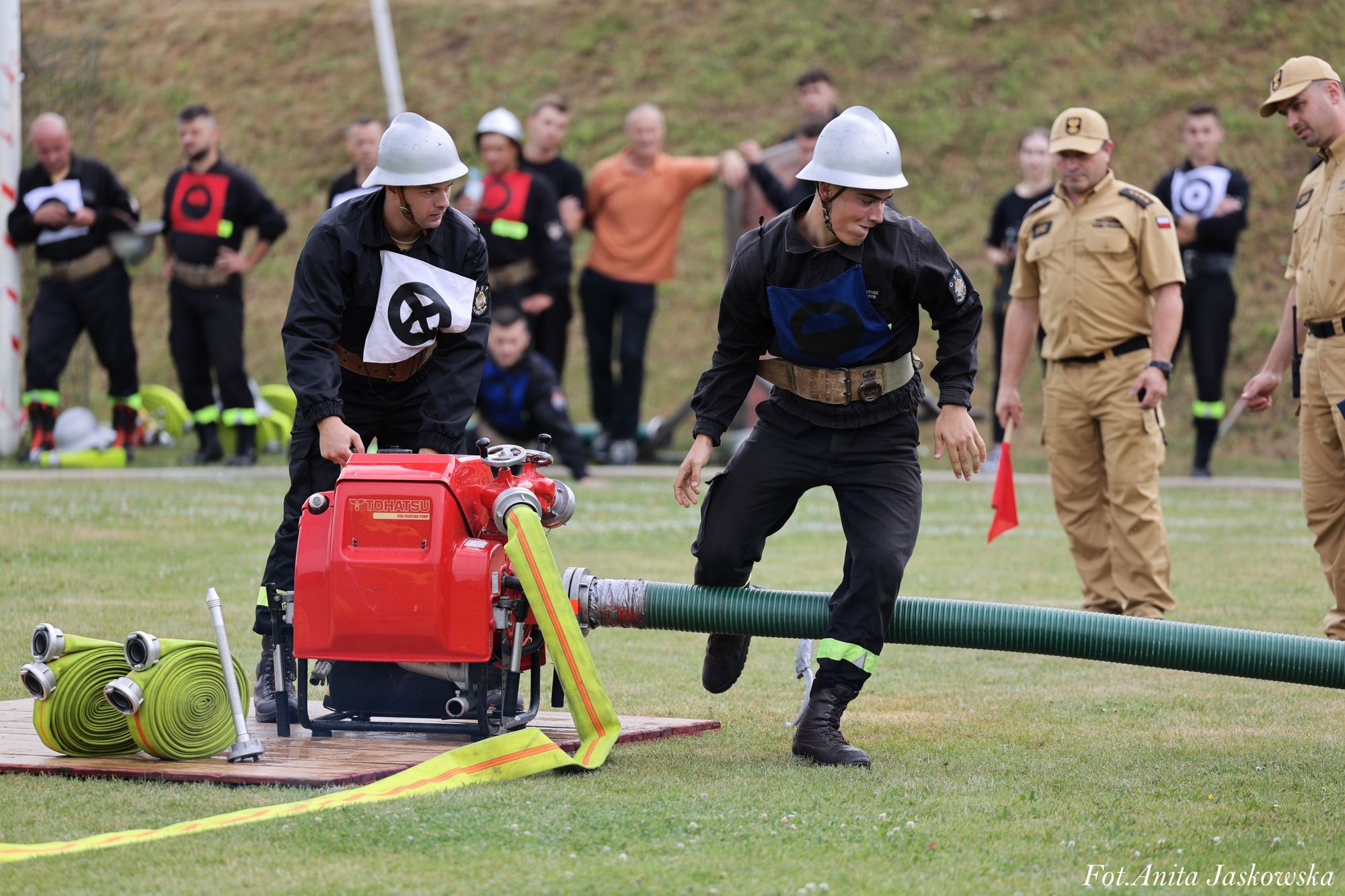 Dwóch strażaków w czarnych mundurach i białych hełmach obsługuje czerwoną pompę z zielonym i żółtym wężem na zielonej trawie, w tle osoby w beżowych mundurach i widzowie na trawiastym zboczu.