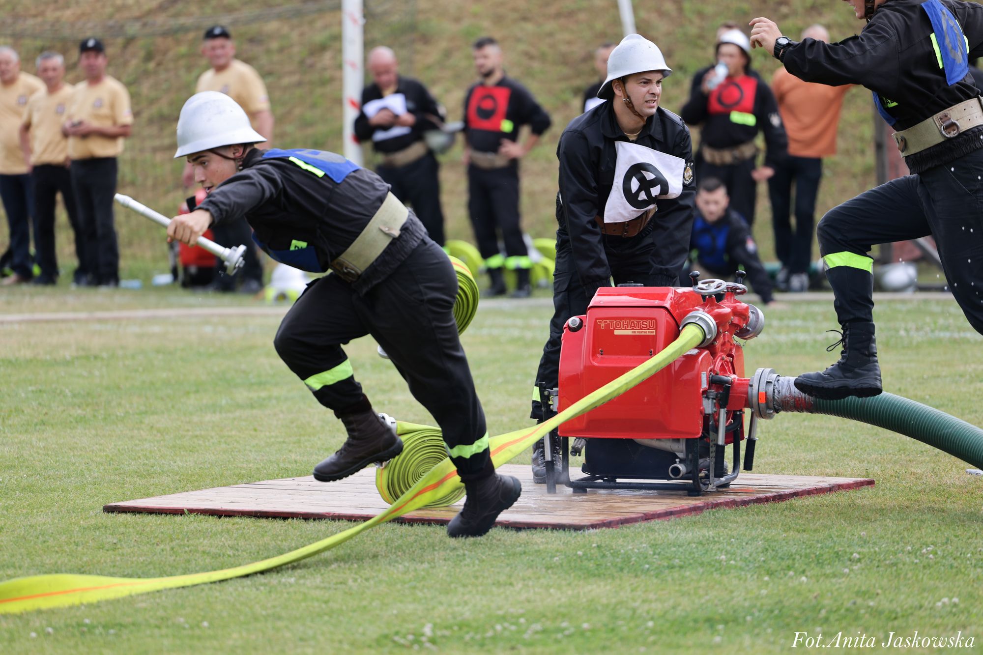 Trzech strażaków w czarnych uniformach i białych hełmach obsługuje czerwoną pompę pożarniczą z podłączonymi żółtymi i zielonymi wężami na trawniku.