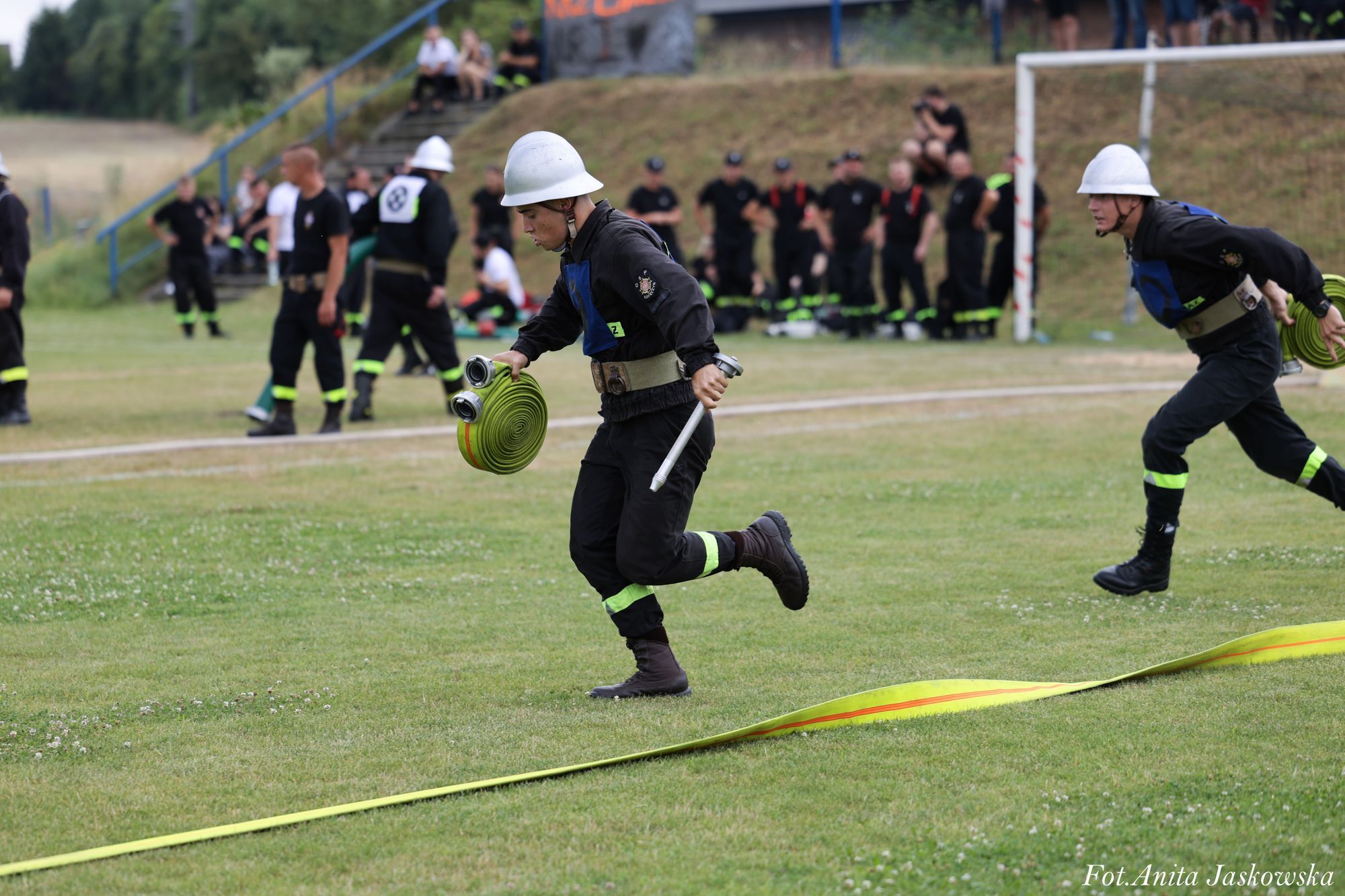 Dwóch mężczyzn w czarnych mundurach z odblaskowymi pasami i białych hełmach biegnie po trawniku, niosąc zwinięte żółte węże strażackie, w tle grupa osób.