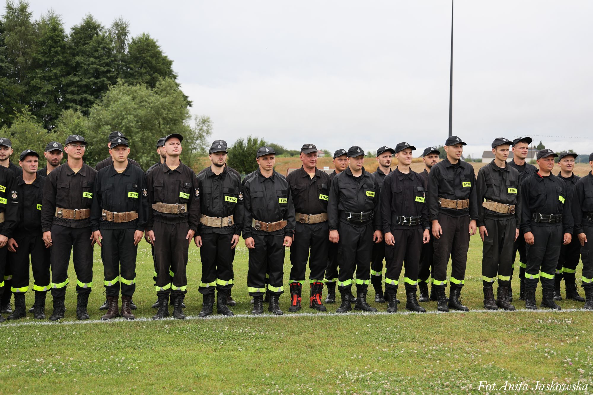 Grupa mężczyzn w czarnych mundurach z neonowymi odblaskowymi pasami na nogawkach i napisami „STRAŻ” na piersiach stoi na zielonej trawie na tle drzew i nieba.