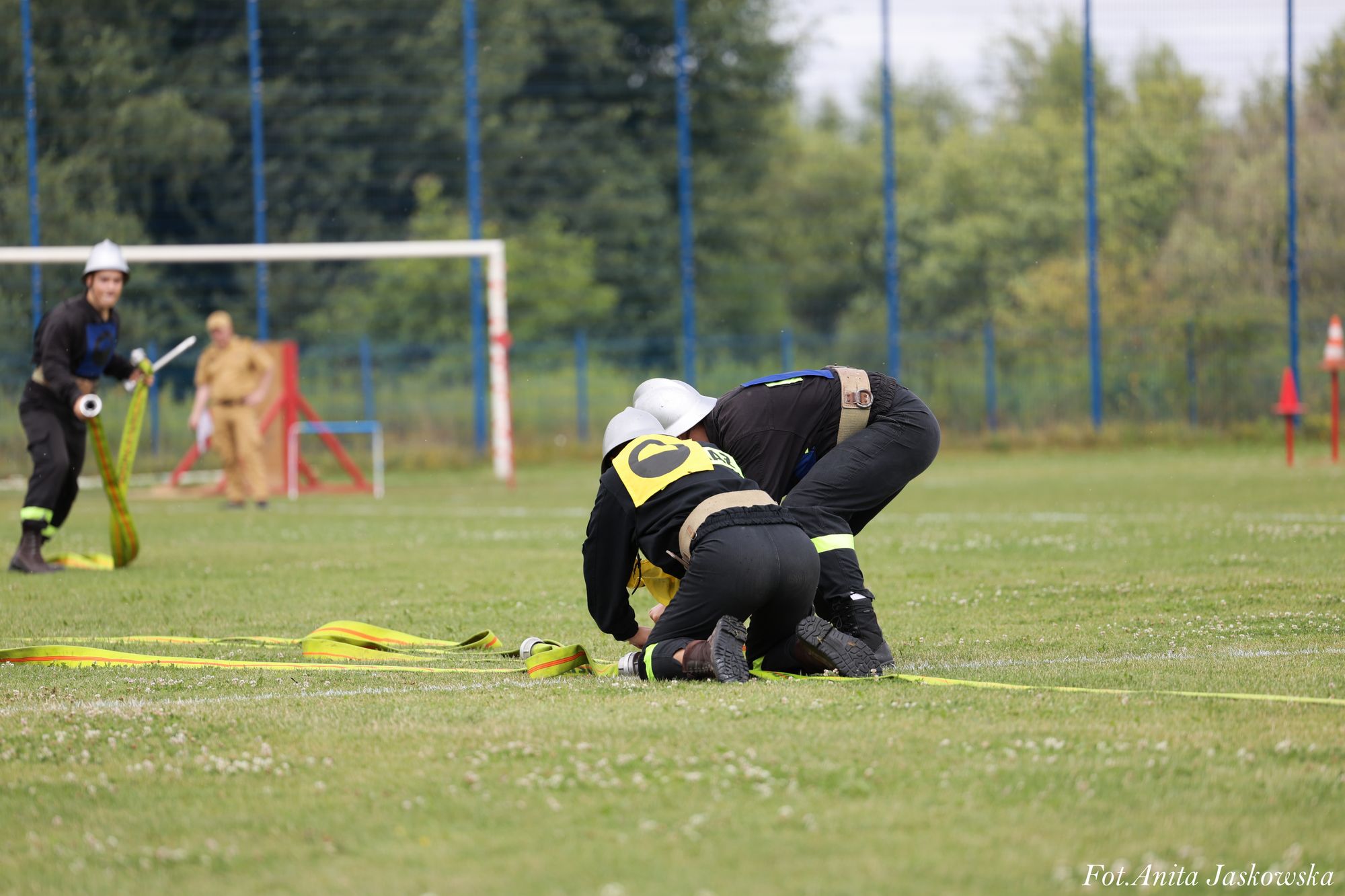 Dwóch strażaków w czarnych mundurach i białych hełmach klęczy na trawie rozwijając żółte węże strażackie, w tle boisko i drzewa.