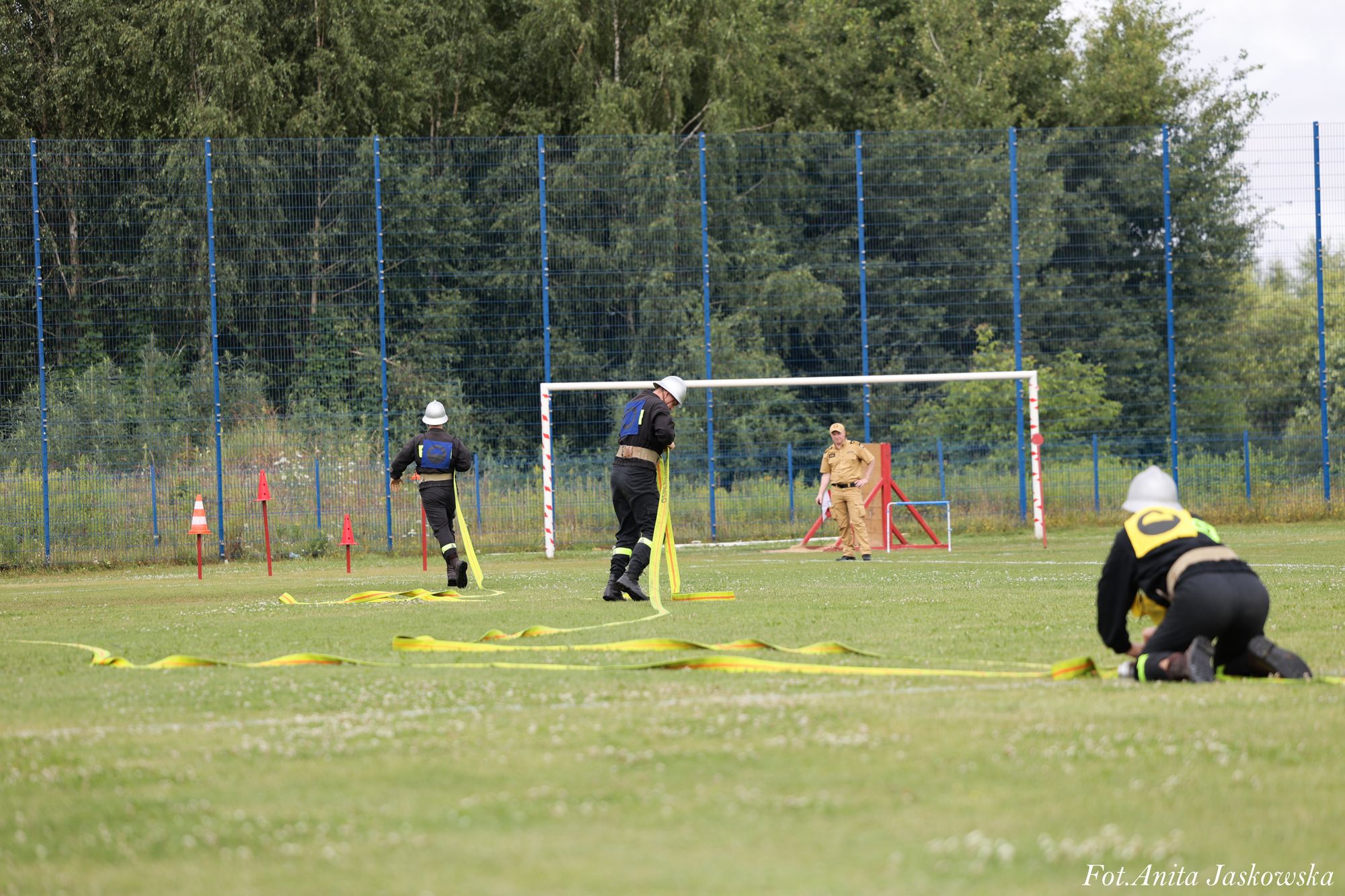 Trzech mężczyzn w czarnych uniformach i białych hełmach z żółtymi taśmami na zielonym boisku, czwarty w beżowym mundurze stoi przy bramce, w tle zielone drzewa i niebieskie ogrodzenie.