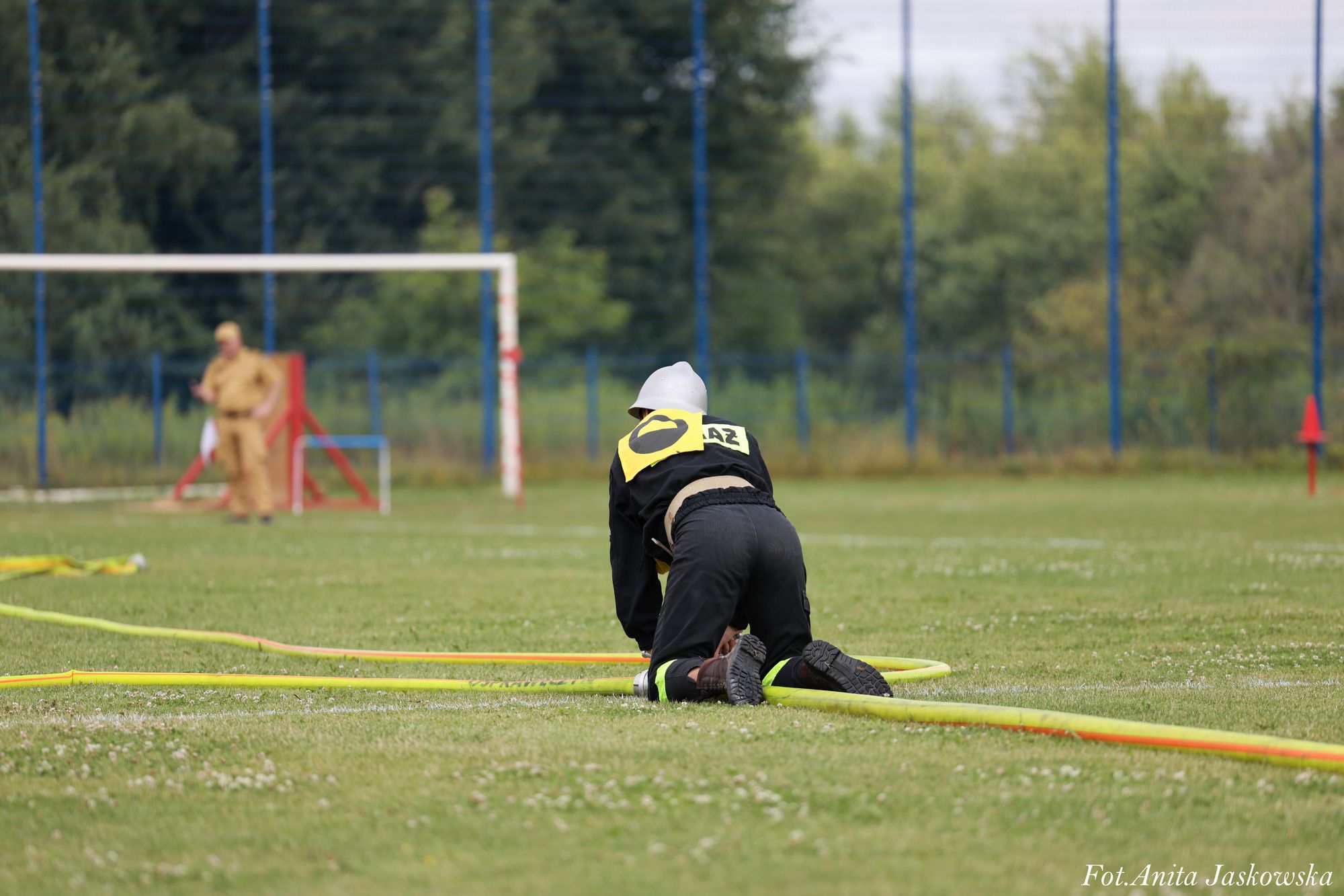 Osoba w czarnym ubraniu ochronnym i białym hełmie klęczy na zielonej trawie obok żółto-pomarańczowego węża strażackiego, w tle bramka i rozmyta postać w brązowym ubraniu.