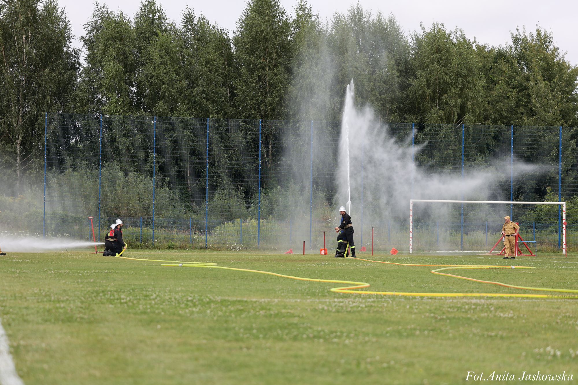 Strażacy w czarnych mundurach i kaskach ćwiczą gaszenie wodą na trawiastym boisku piłkarskim z bramką, w tle zielone drzewa i niebieska siatka ogrodzenia.
