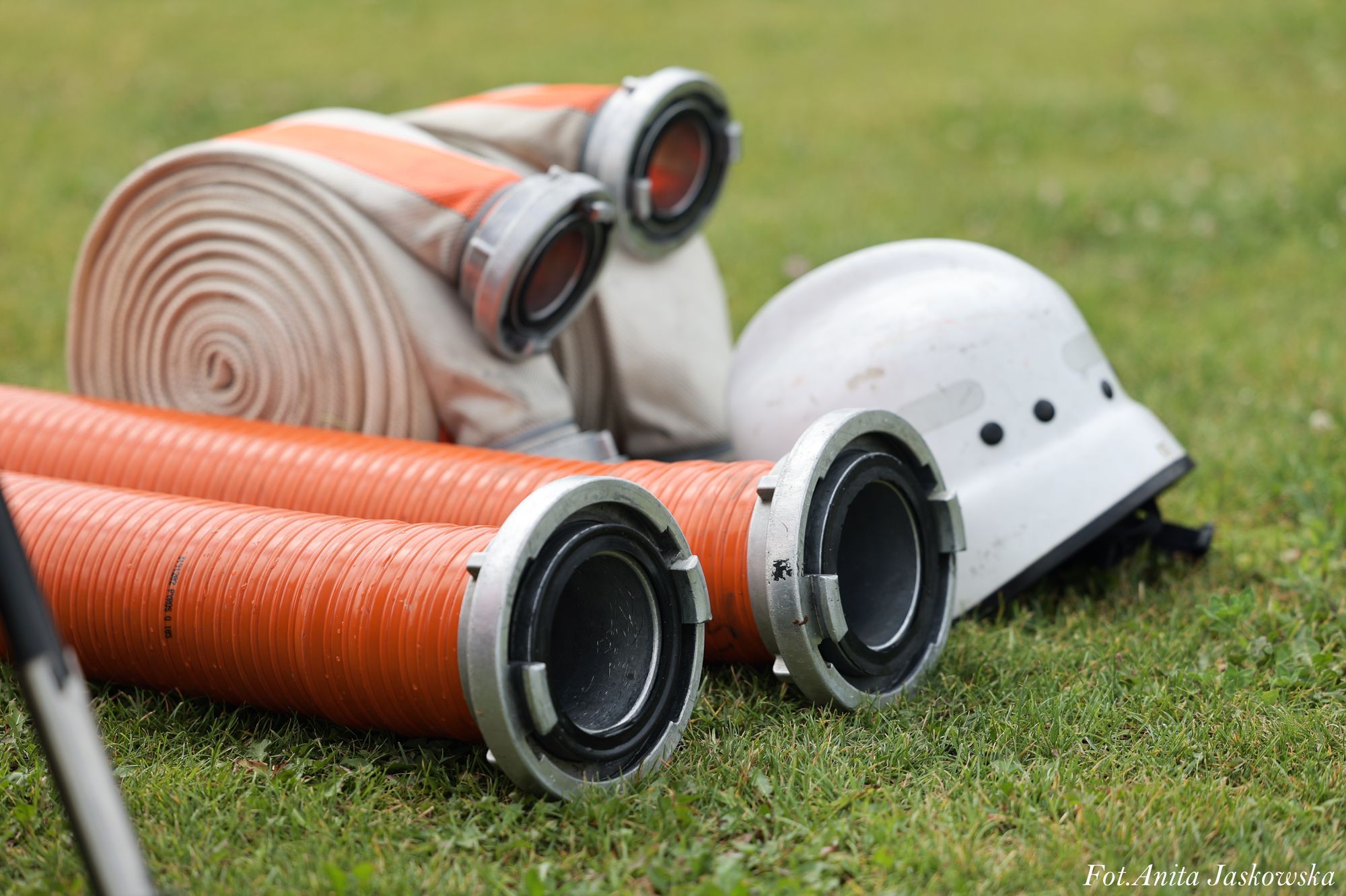 Dwa pomarańczowe węże pożarnicze z metalowymi końcówkami, zwinięty beżowy wąż oraz biały hełm na zielonej trawie.