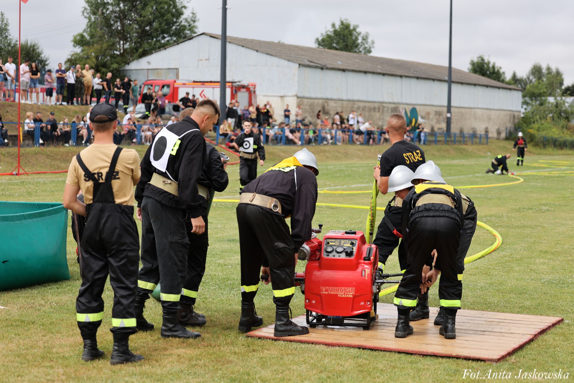 Strażacy w czarnych mundurach i srebrnych hełmach obsługują czerwoną pompę wodną na drewnianej platformie na trawiastym boisku, w tle widownia i budynek gospodarczy.