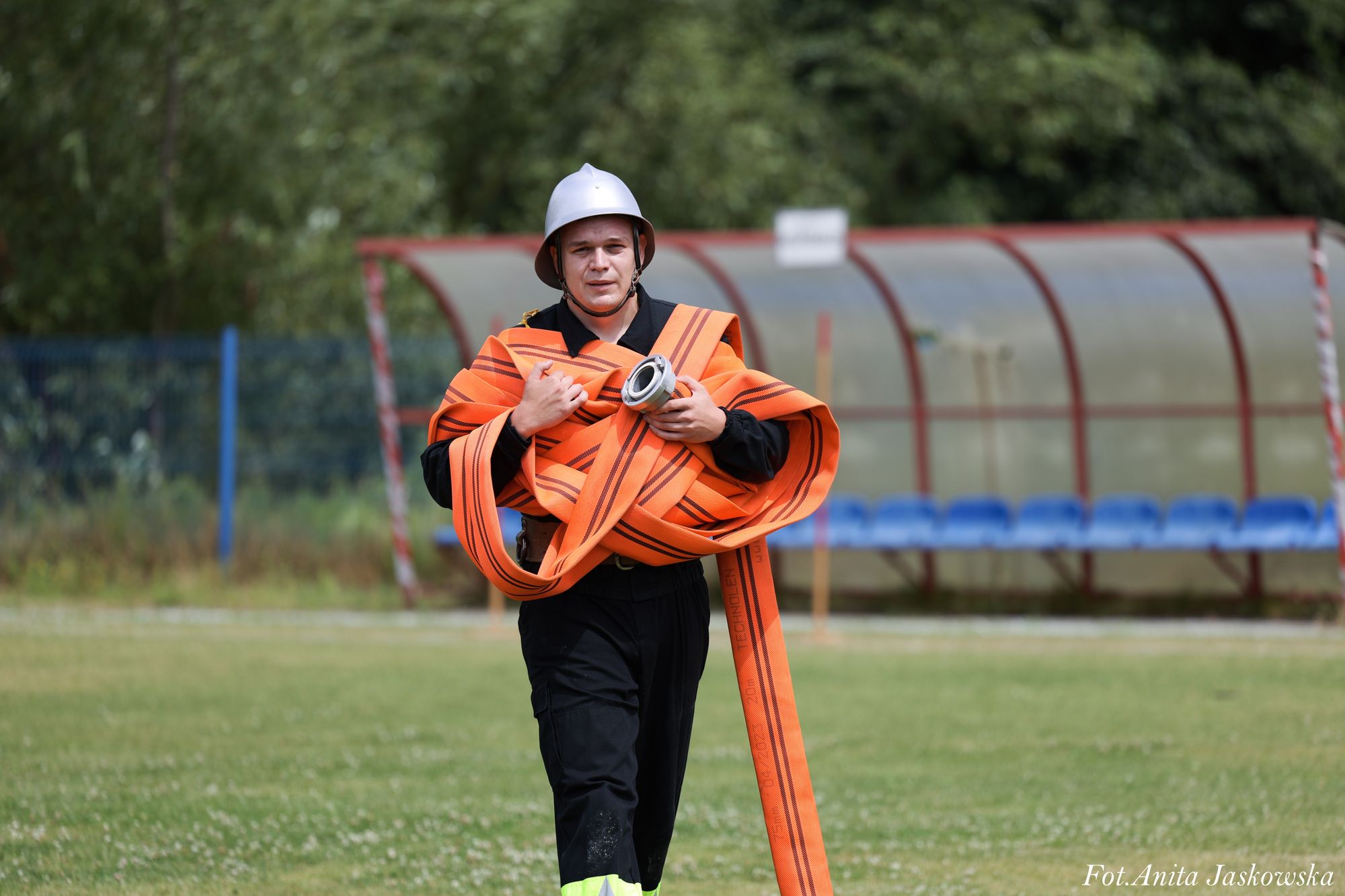 Mężczyzna w czarnym ubraniu i srebrnym hełmie niesie zwinięty pomarańczowy wąż strażacki na zielonym boisku, w tle zadaszone niebieskie ławki.
