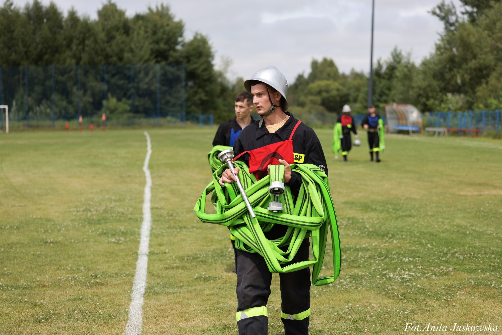 Mężczyzna w hełmie i ciemnym ubraniu z czerwonym elementem niesie zielony zwinięty wąż strażacki na tle trawiastego boiska i drzew. W tle inni ludzie w podobnych strojach.
